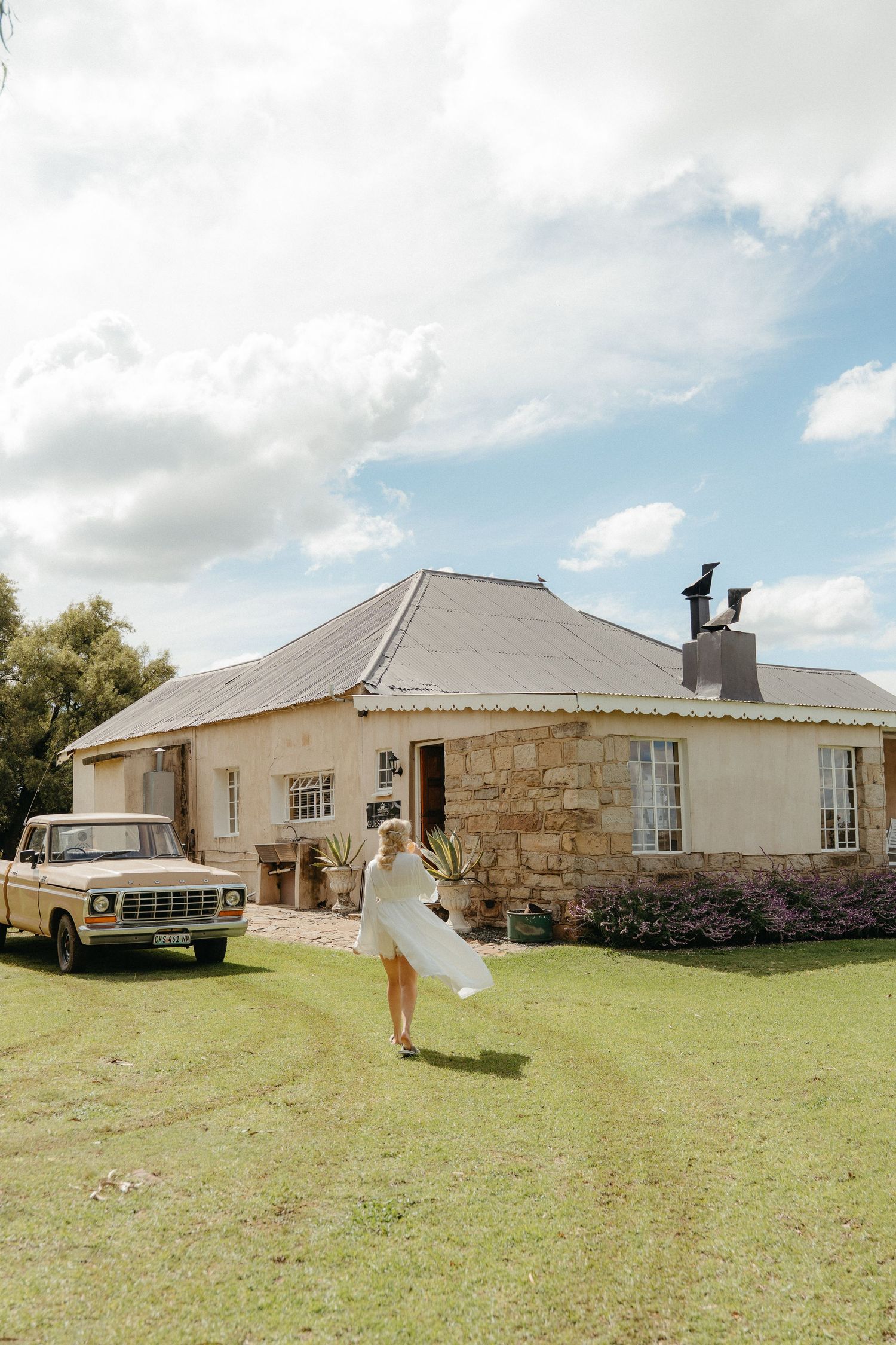 Bride in white dress walking toward rustic stone farmhouse with vintage truck on green lawn under cloudy sky.