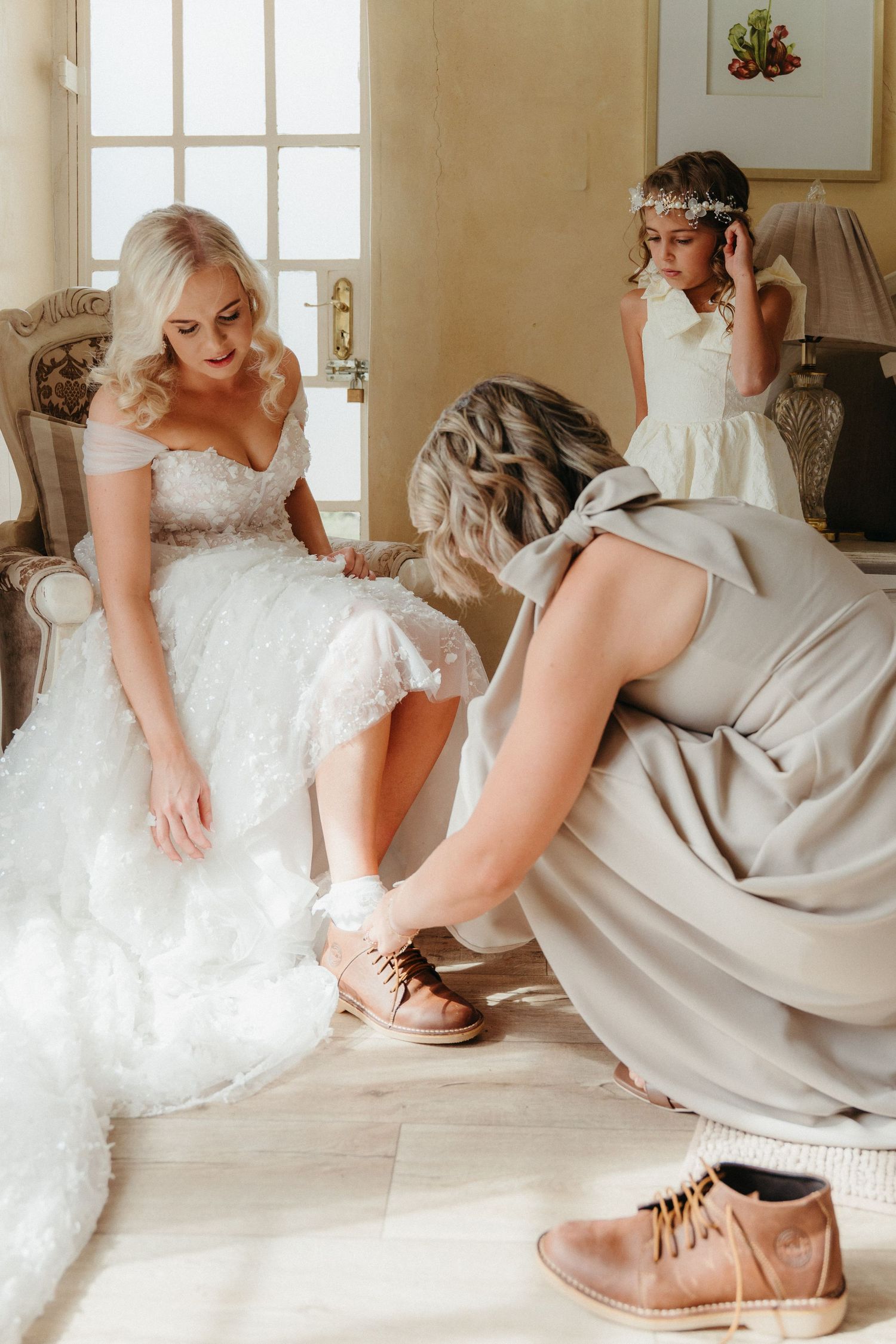 Bridesmaid helping bride put on brown boots while flower girl watches during wedding preparations indoors.