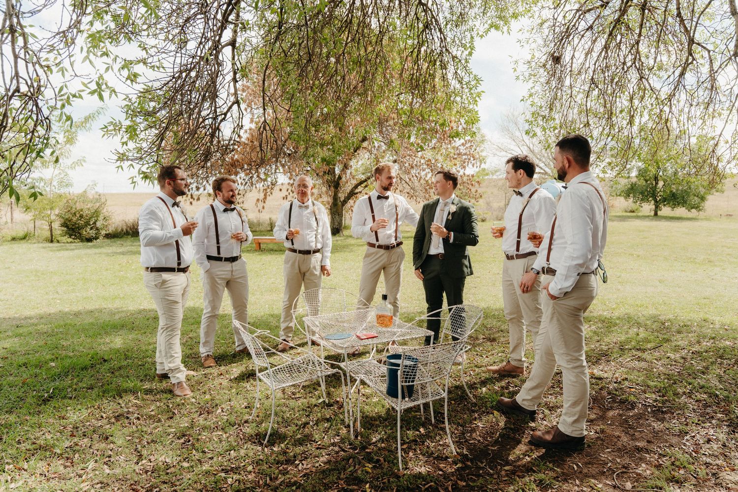Groomsmen in cream suits socializing outdoors on a sunny day near a white iron table.