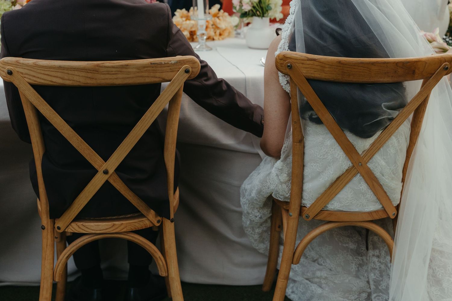Bride and groom sitting together at wedding reception table on elegant wooden cross-back chairs, holding hands.