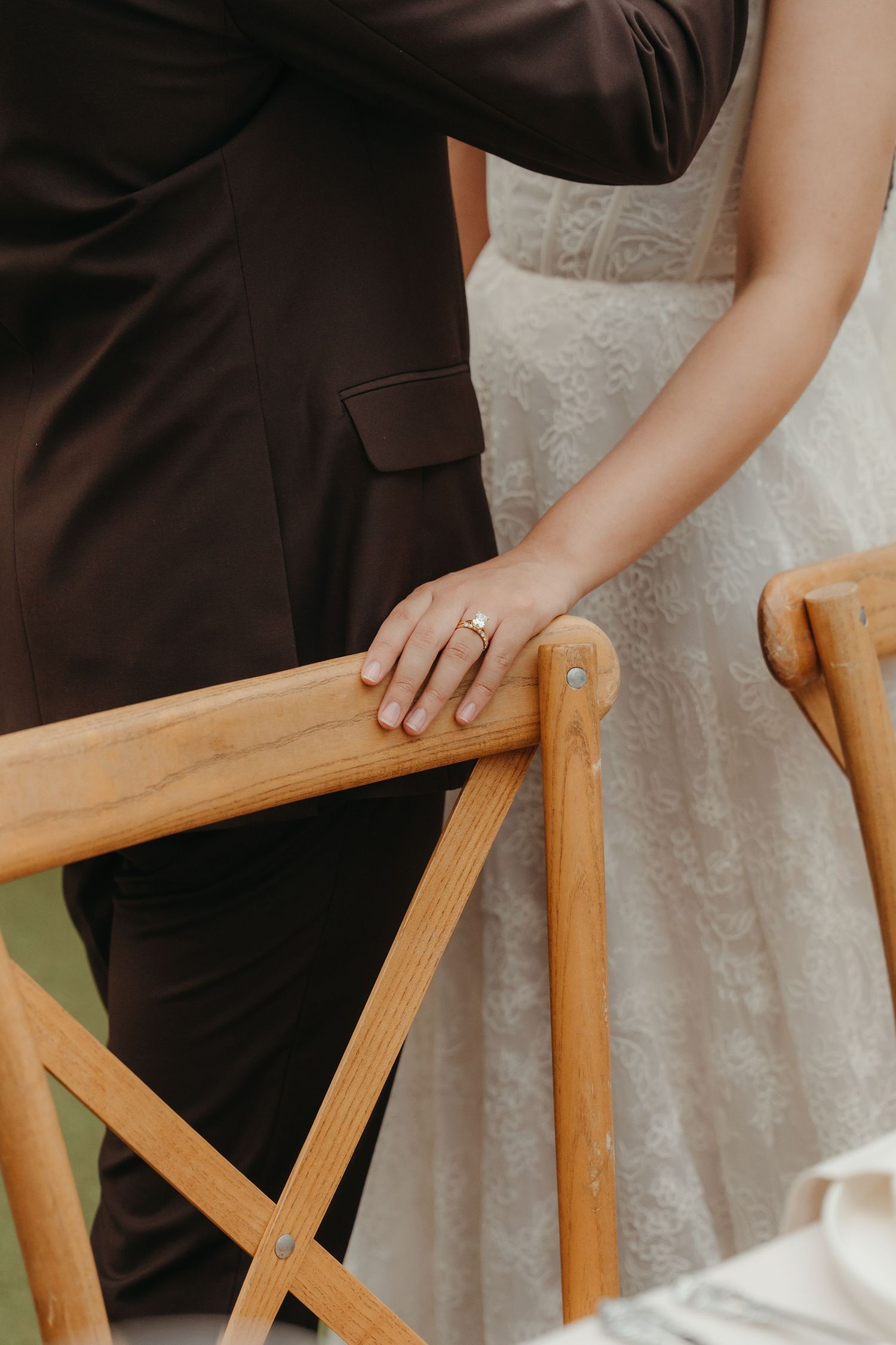 Bride and groom standing together, her hand resting on a wooden chair, showcasing a sparkling wedding ring.