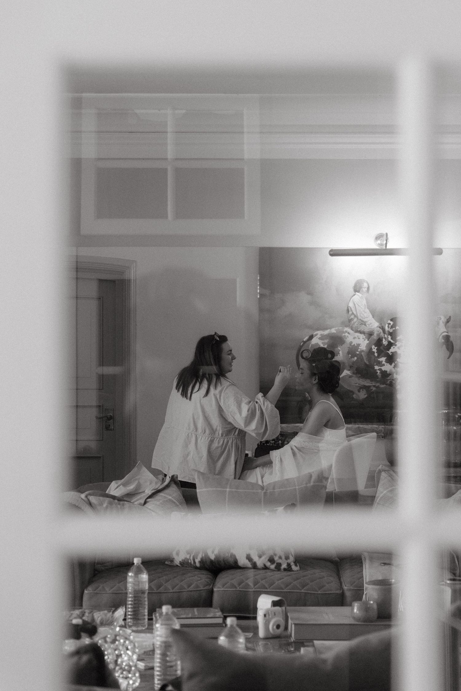 Black and white photo of a bride getting ready, seen through a window, with a mural visible in the background.