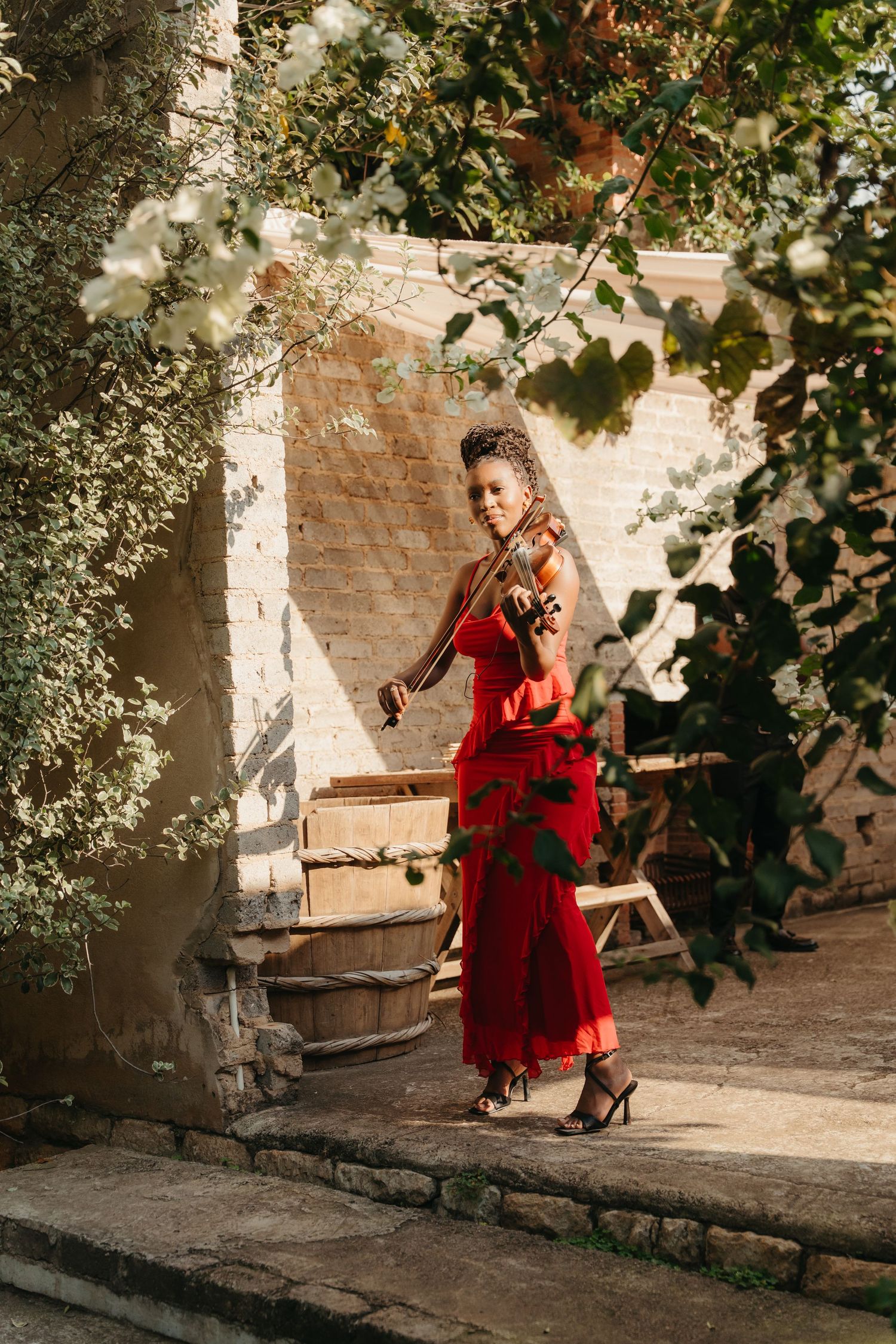 Woman in red dress playing violin outdoors, surrounded by lush green foliage and rustic stone architecture.