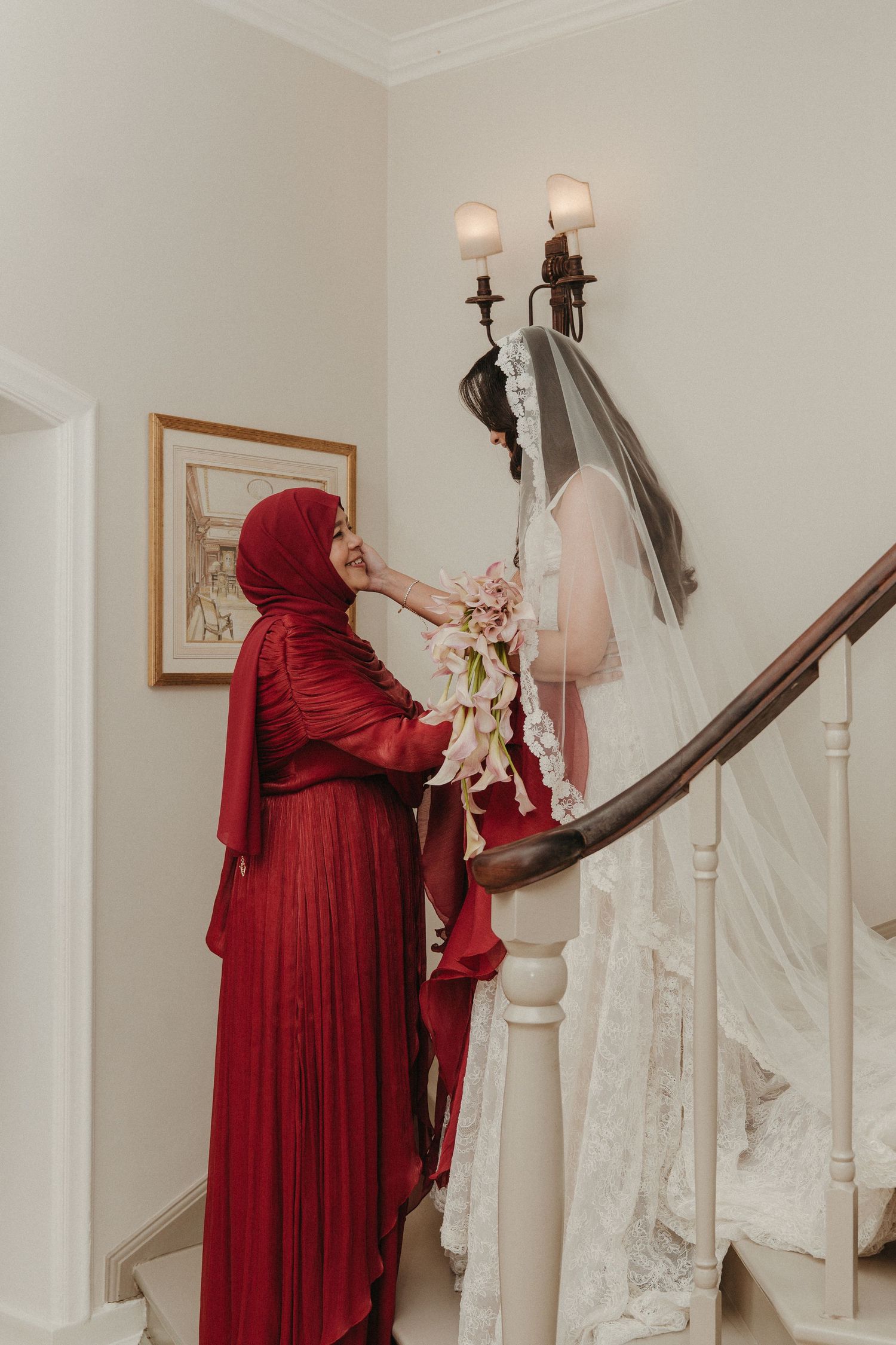 A woman in a red hijab helps a bride in a white lace gown and veil on an elegant staircase.