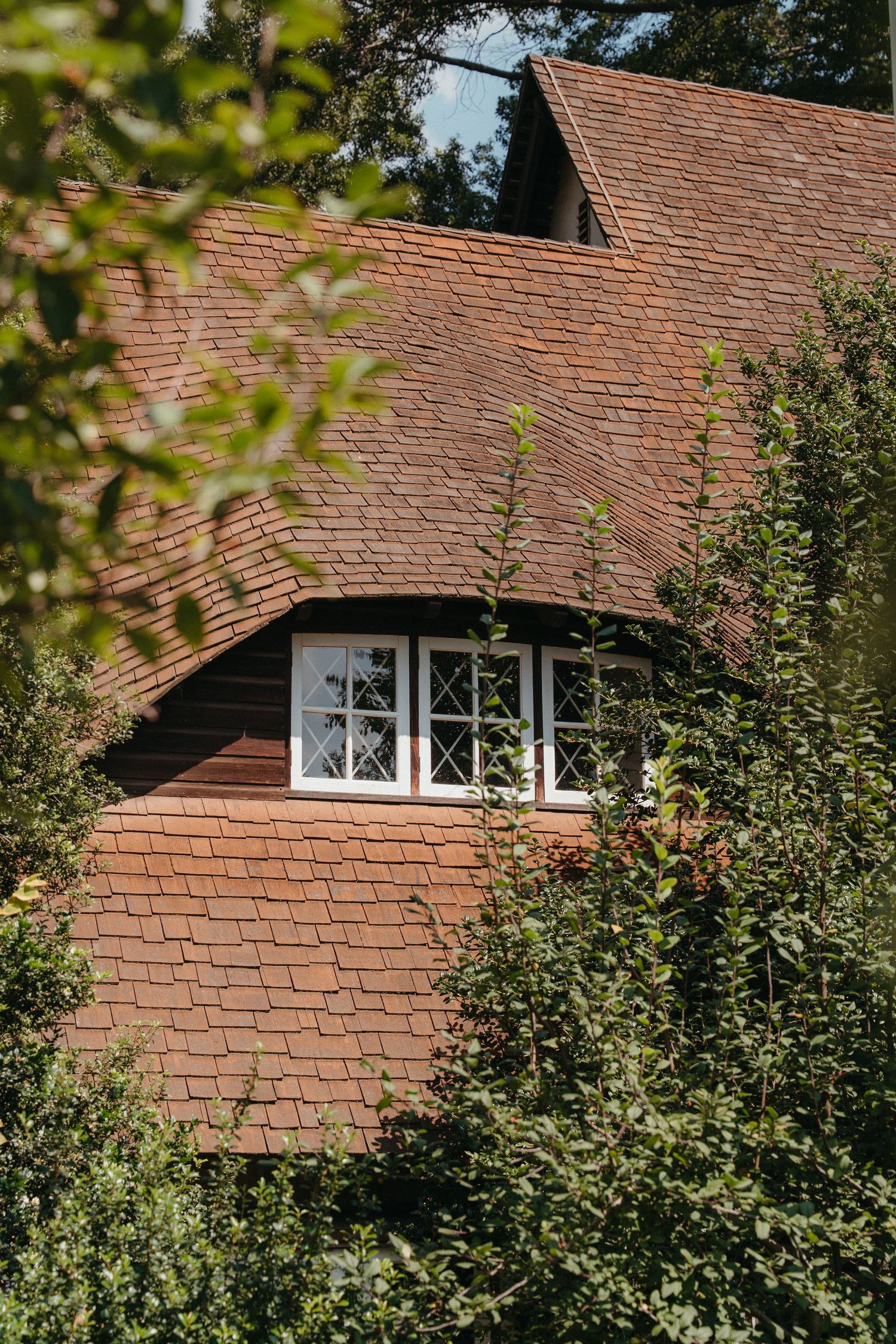 A charming red-brick cottage with white-framed windows, steep tiled roof, surrounded by lush green trees and climbing plants.
