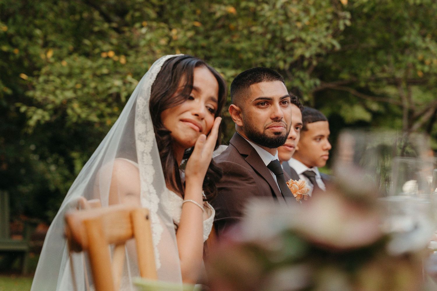 Bride in veil smiles emotionally at outdoor wedding reception, seated beside groom in dark suit.