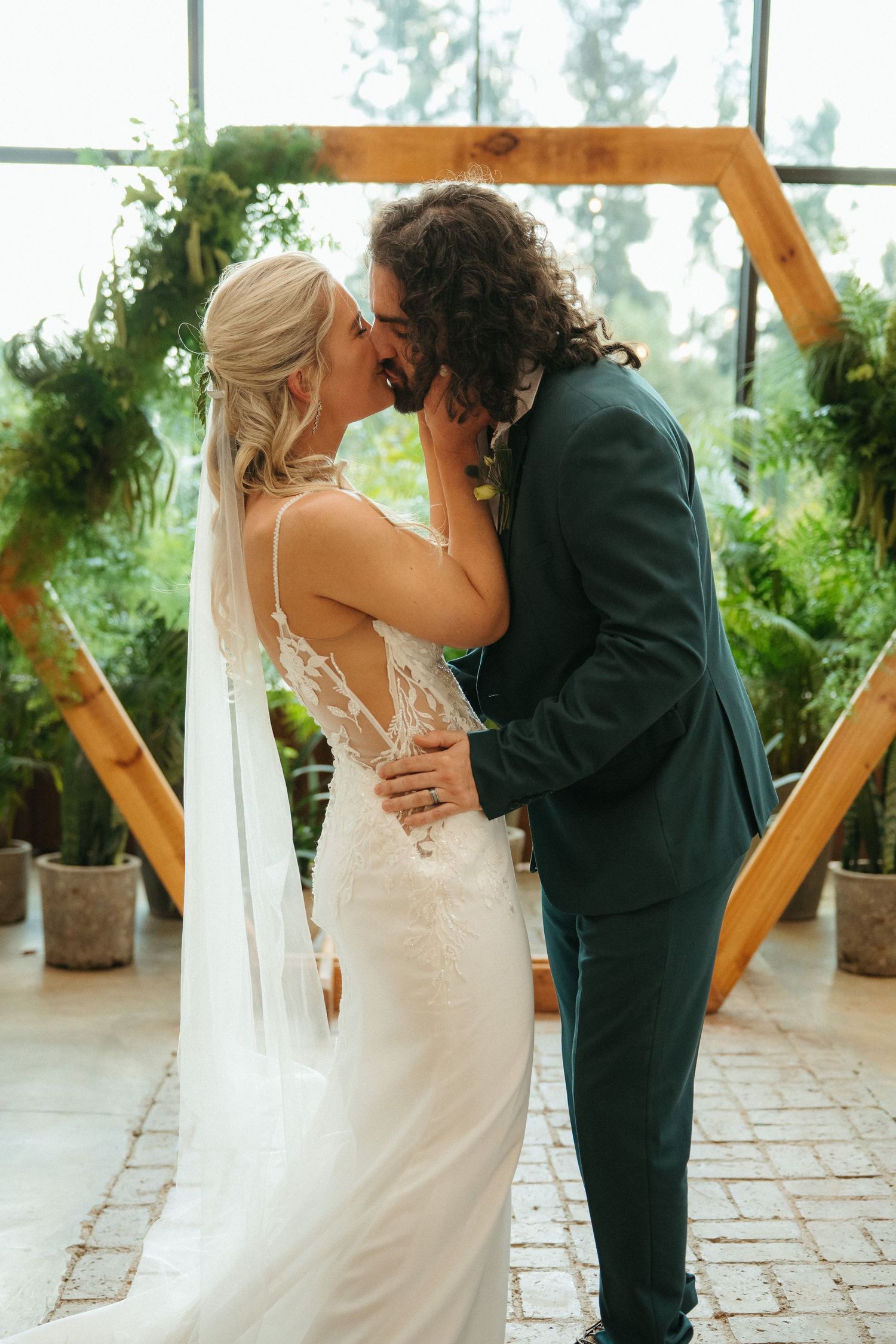 Bride and groom share first kiss at botanical wedding venue with hexagonal wooden arch and lush greenery backdrop.