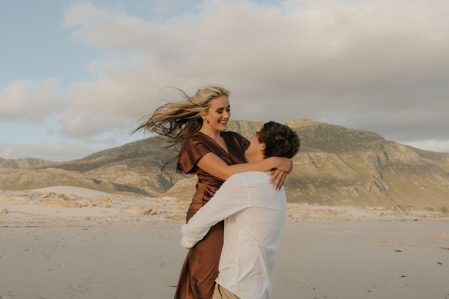 Couple sharing a joyful embrace on a sandy beach with mountains and cloudy sky in the background.