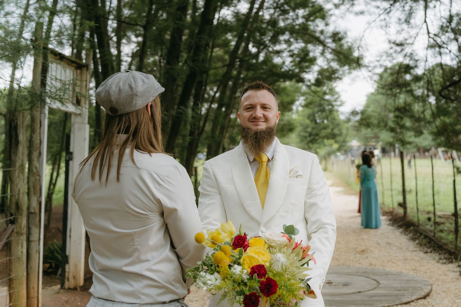 Wedding couple exchanging vows on a tree-lined path with yellow and red floral bouquet.
