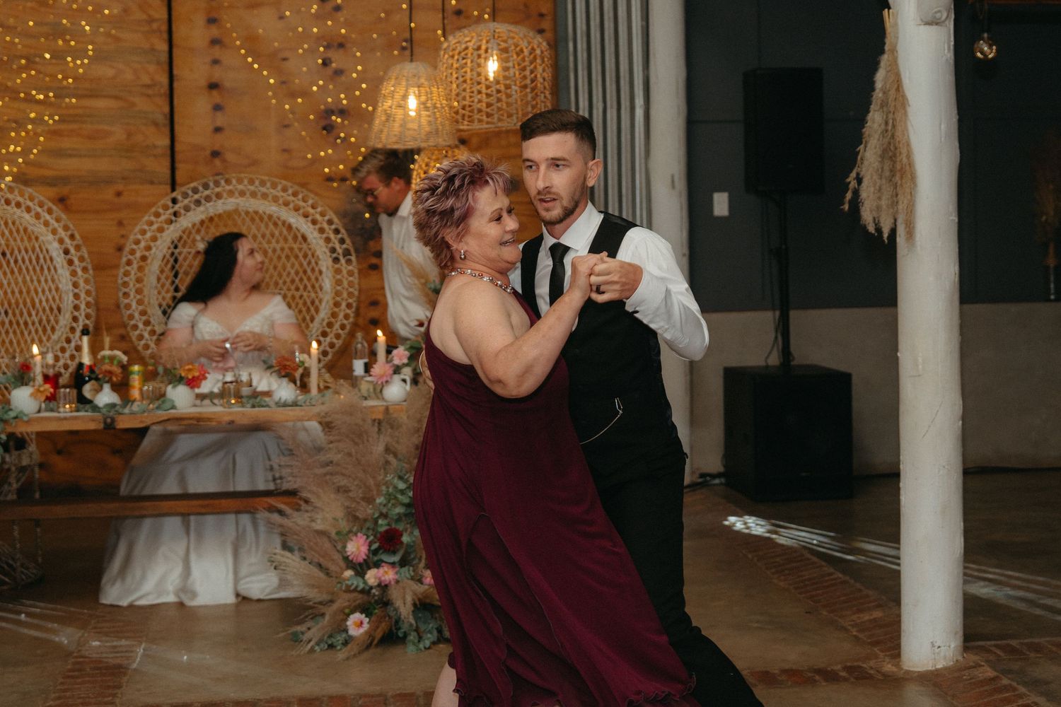 A couple shares a romantic dance together at their wedding reception with decorative lighting and peacock chairs in the background.