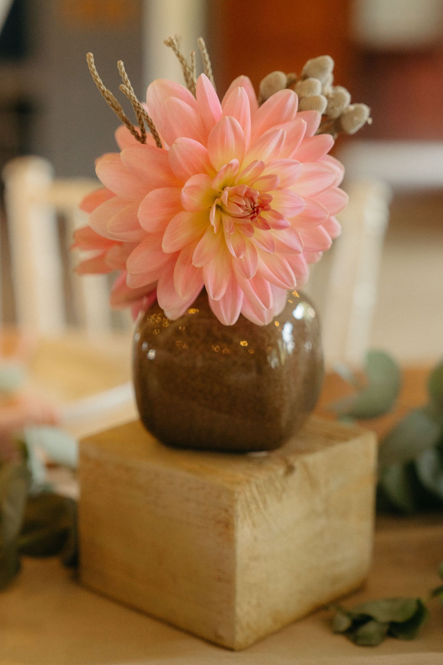 A peachy pink dahlia flower in a brown glass vase sits atop a wooden block with greenery accents.