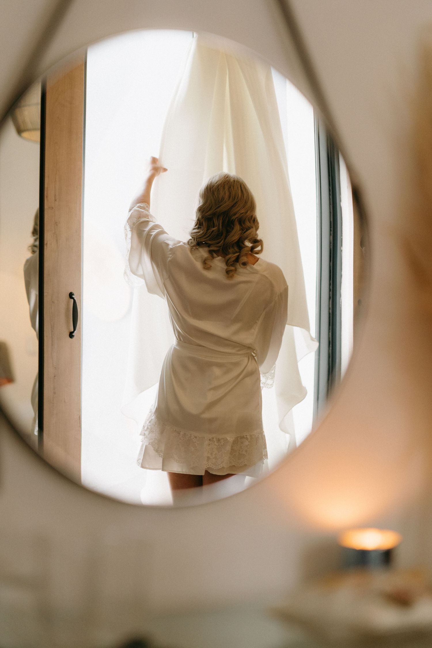 A figure in a white robe looking out through curtained windows, reflected in a circular mirror with soft lighting.