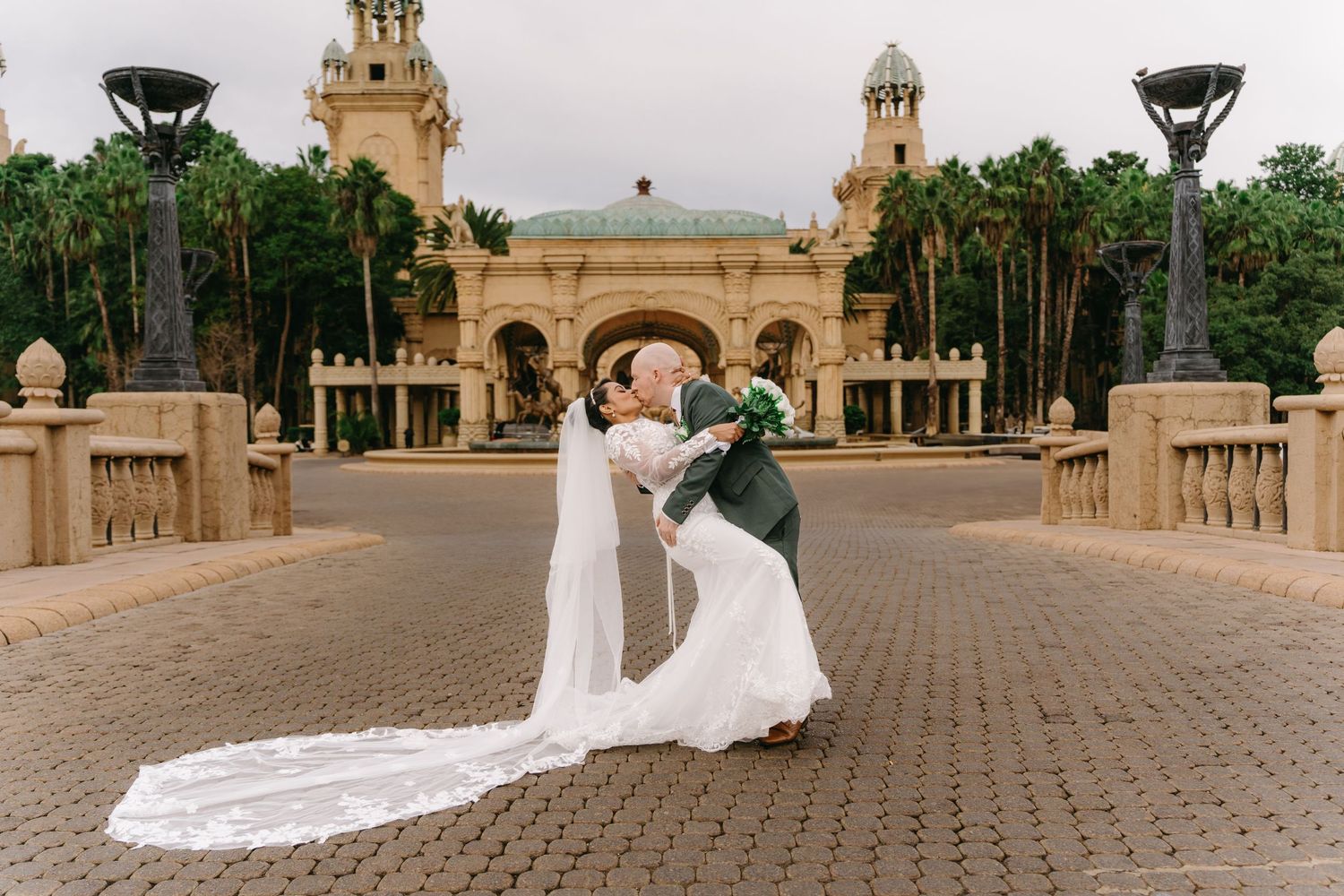 A couple in wedding attire poses on a grand staircase at a luxurious Mediterranean-style venue with ornate architecture.