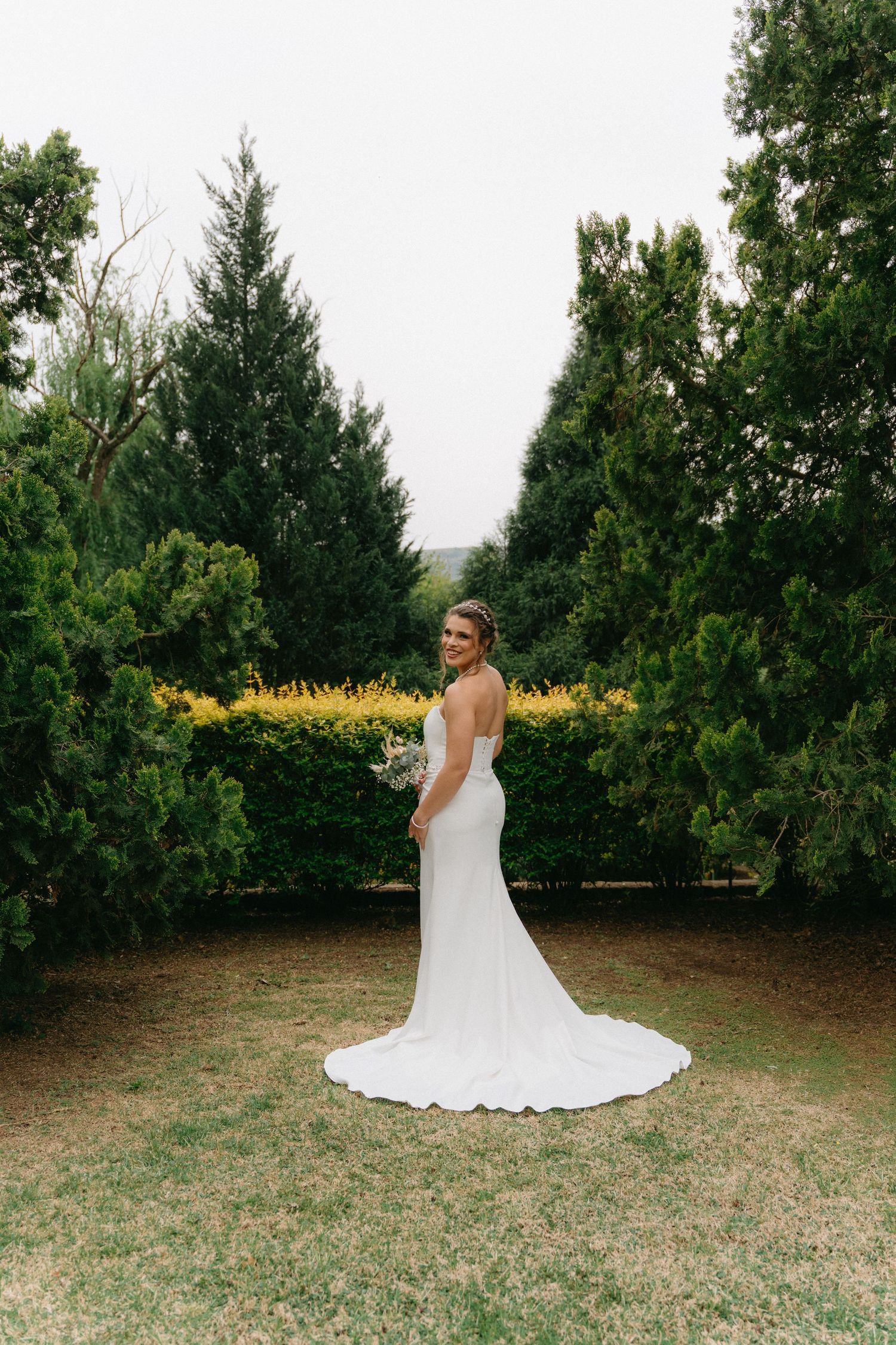 Elegant white wedding dress photographed in garden setting with evergreen trees in background.