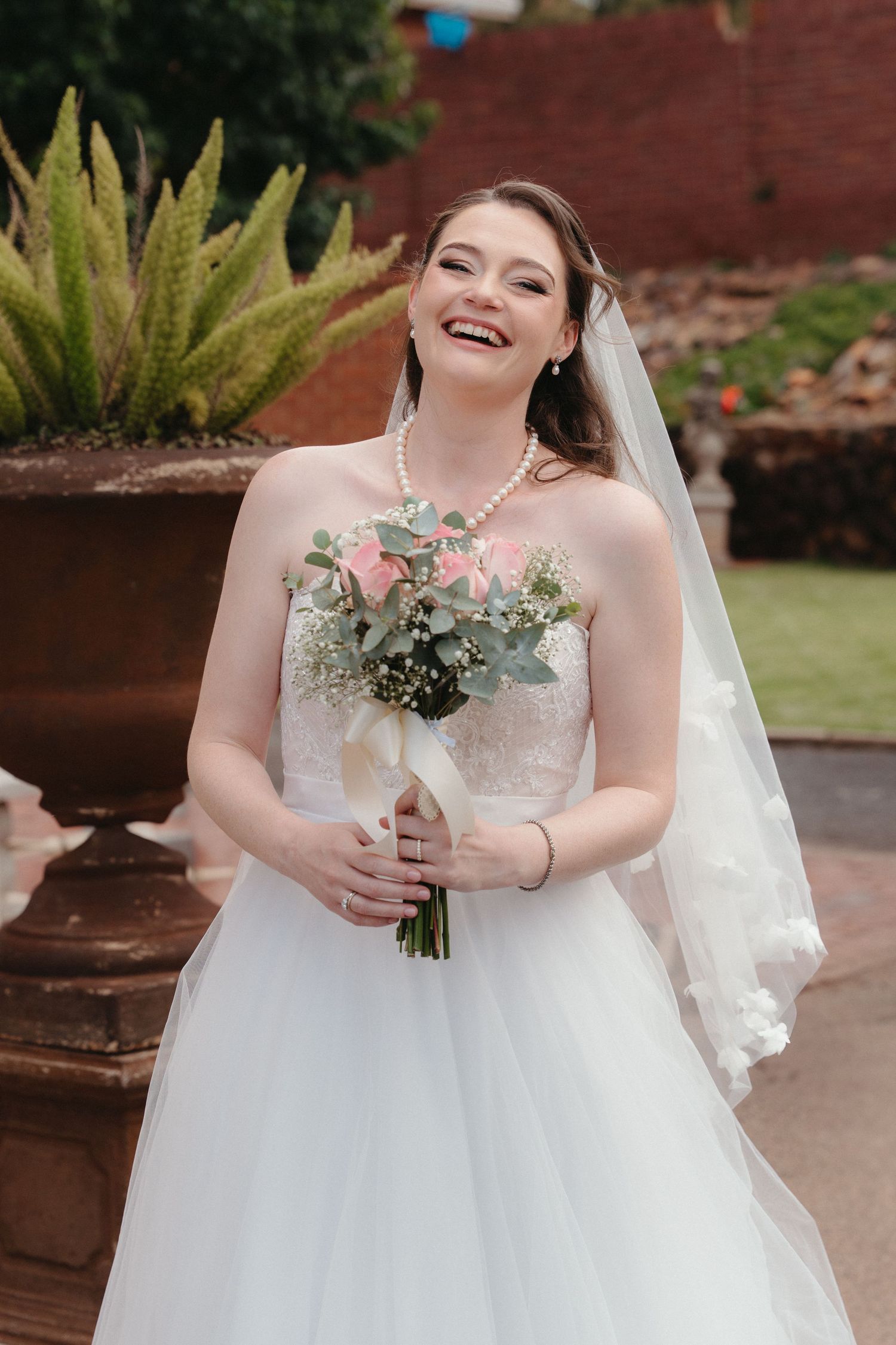 A bride in a white wedding gown holds a delicate bouquet of flowers and greenery against a brick backdrop at Nazareth House Pretoria