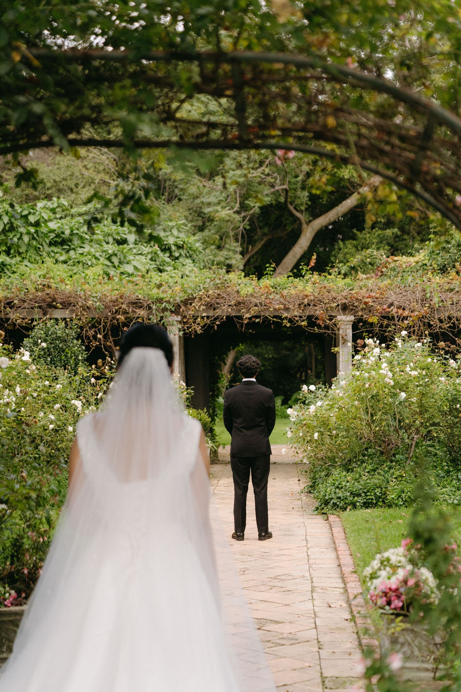 A bride in a flowing white gown approaches her groom for their first look in a lush garden pathway.