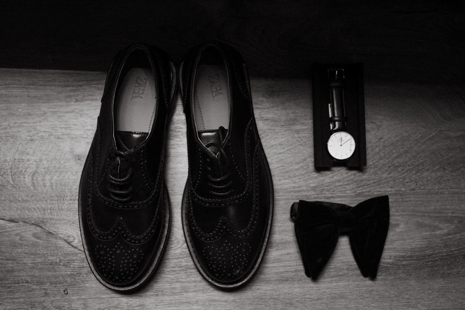 Black dress shoes, bowtie, and accessories arranged on wooden floor for formal event.