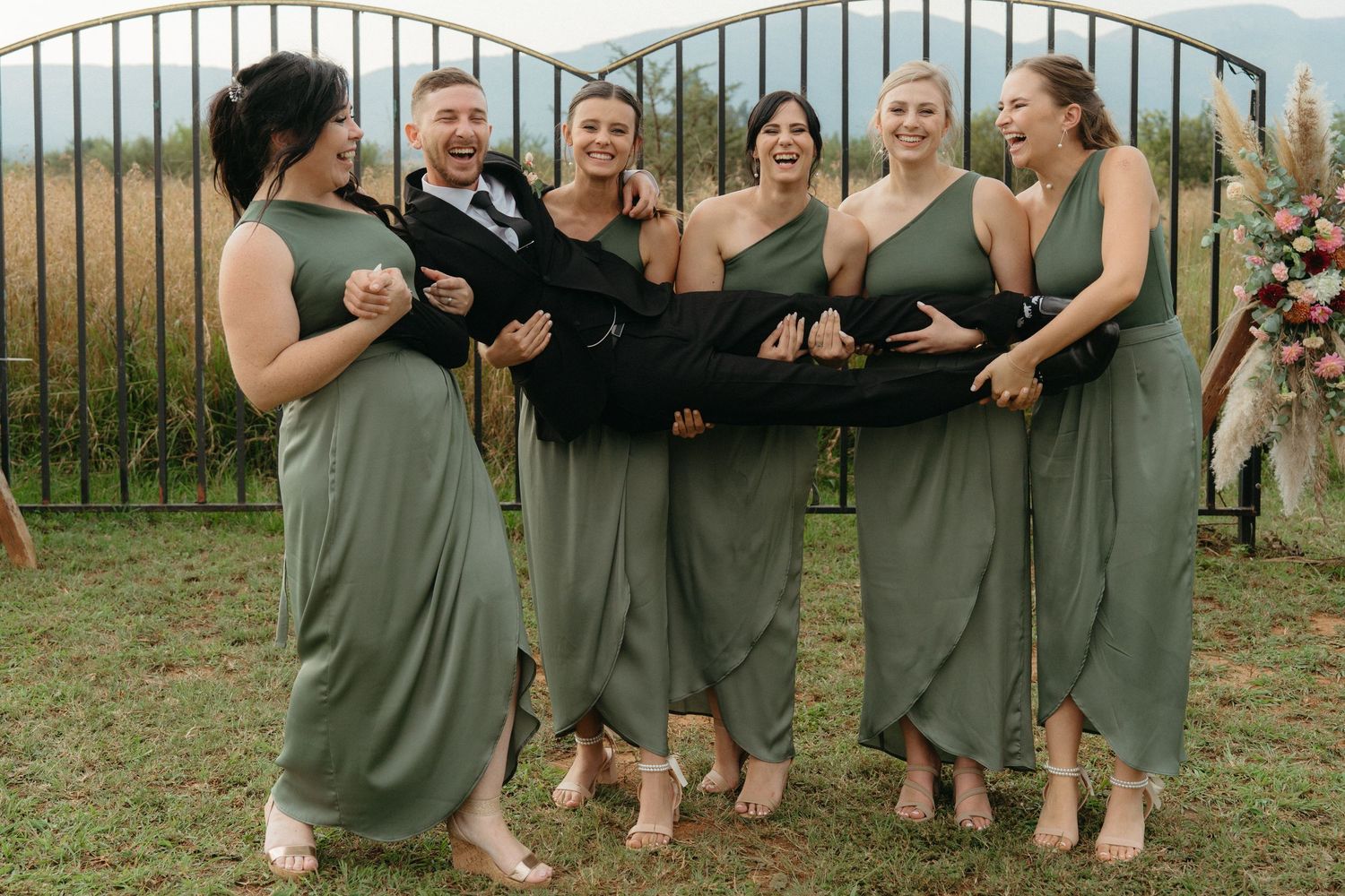 A group of bridesmaids wearing sage green dresses stand together laughing on a grassy lawn behind an iron fence.
