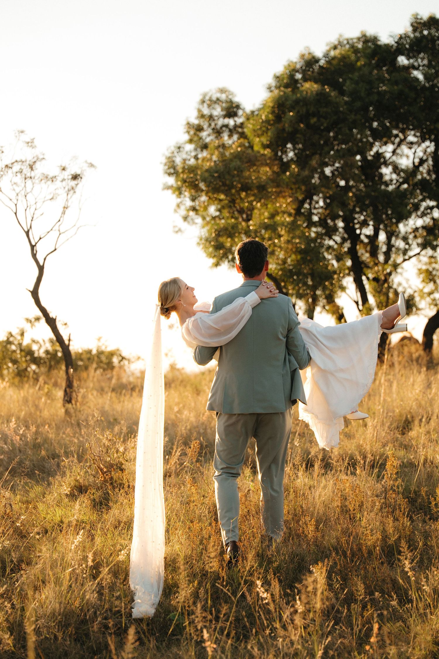 Groom carrying bride in white gown through bushveld field at sunset in Rustenburg