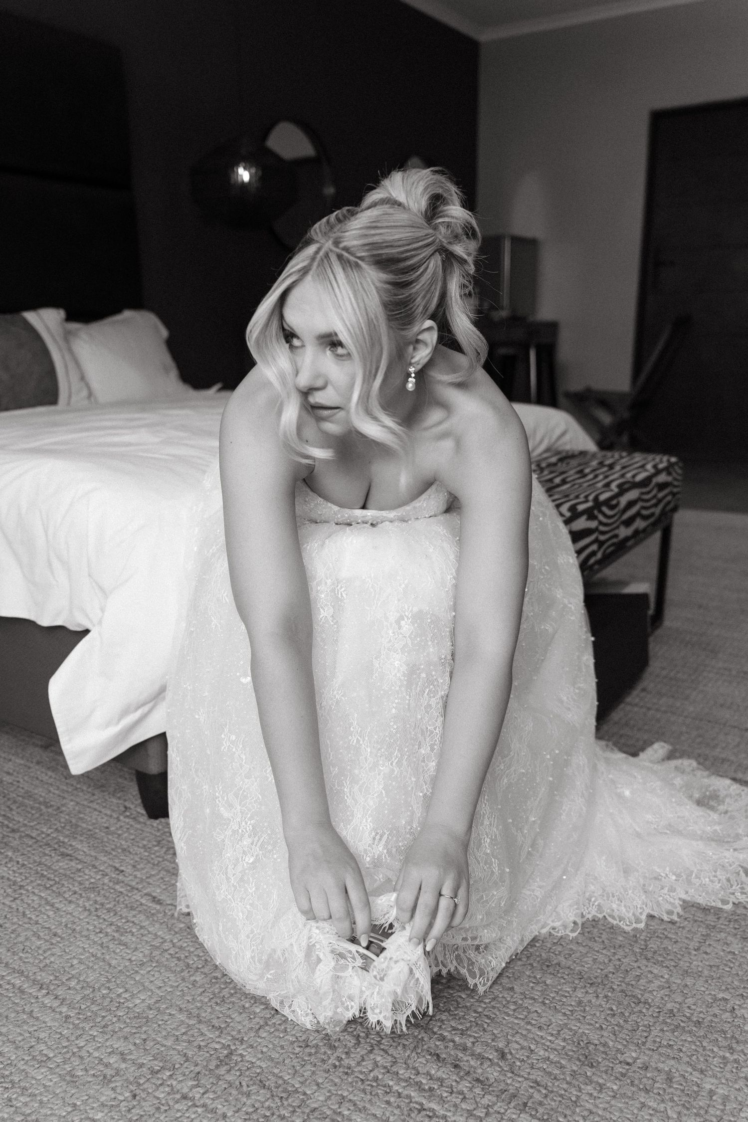 Black and white photo of blonde bride in lace gown sitting on floor adjusting shoes by bed at Simbali Lodge in Pretoria