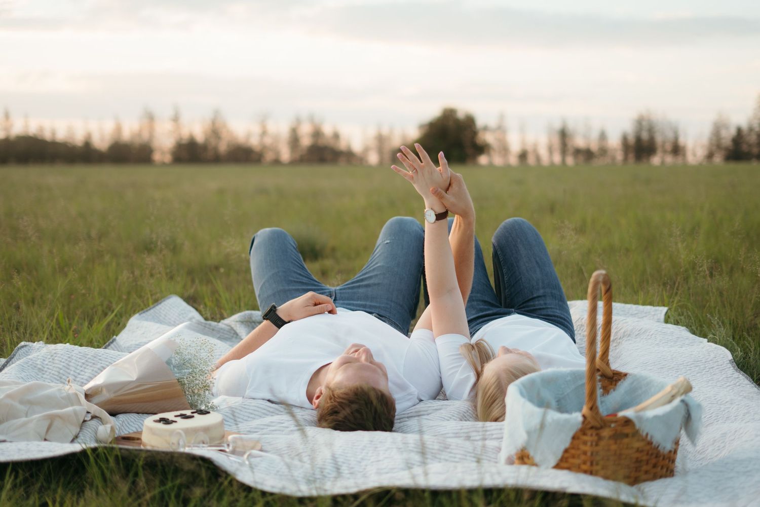 Couple lying on picnic blanket in grassy field reaching toward sky.