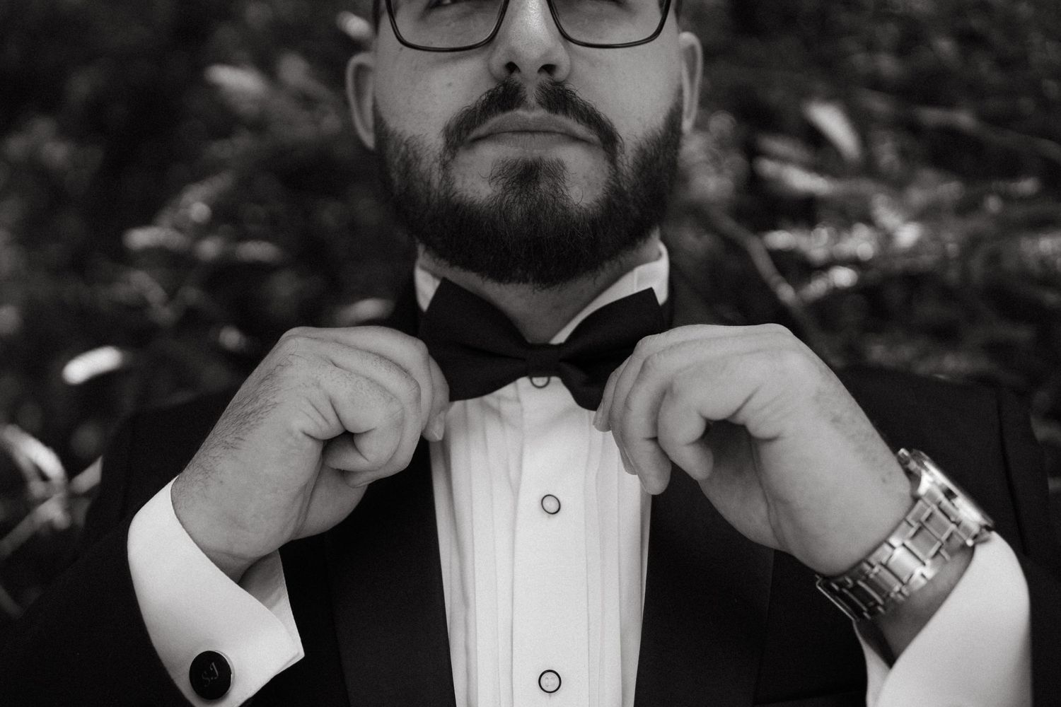 Person in formal attire adjusts bowtie while wearing glasses and wristwatch in black and white photo.