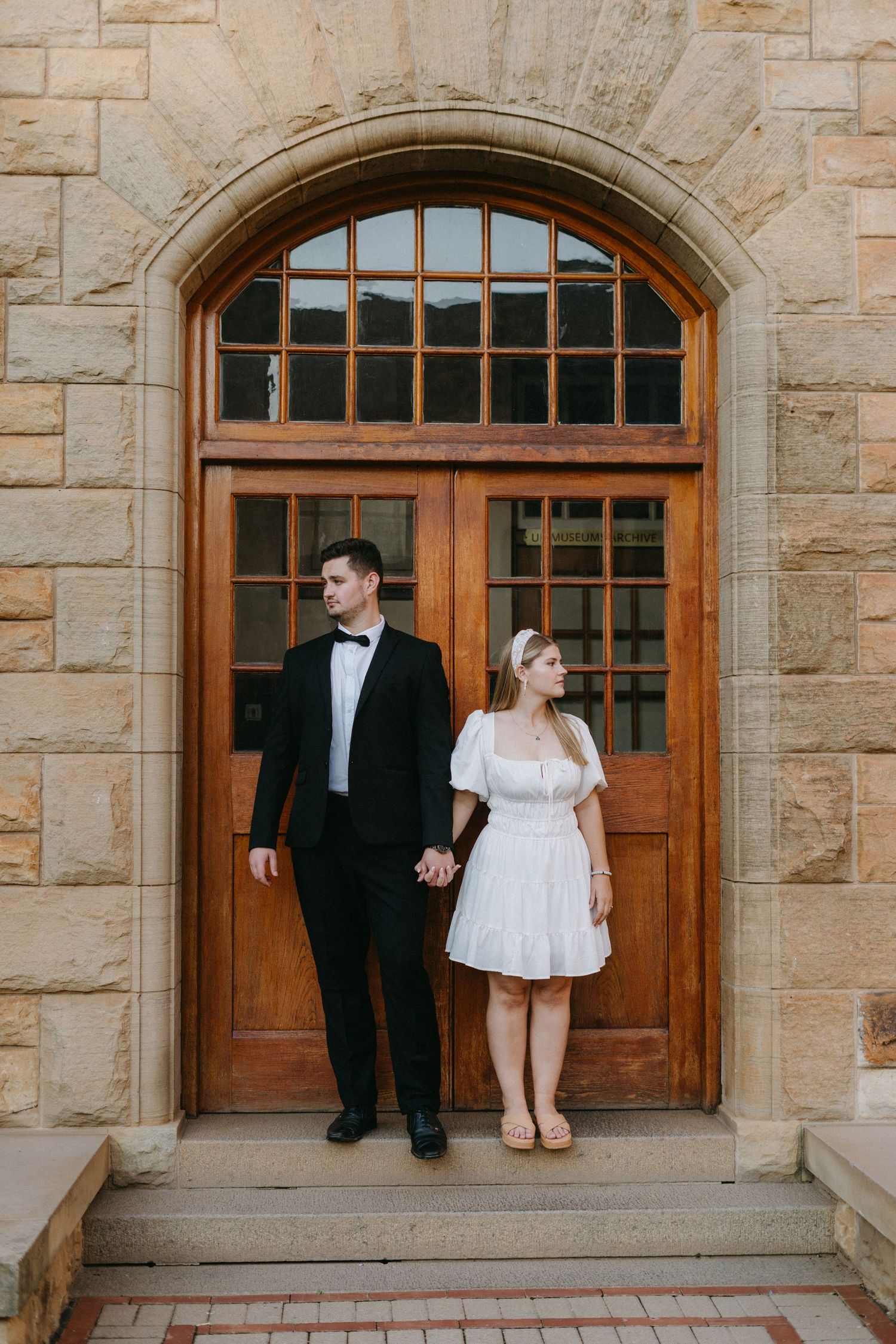 Newlywed couple in formal attire standing together by an arched wooden doorway in a stone building.