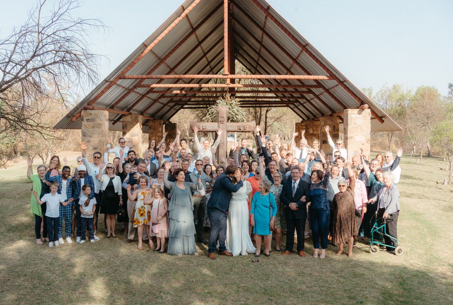 A large group celebrates under a rustic wooden pavilion at an outdoor event.
