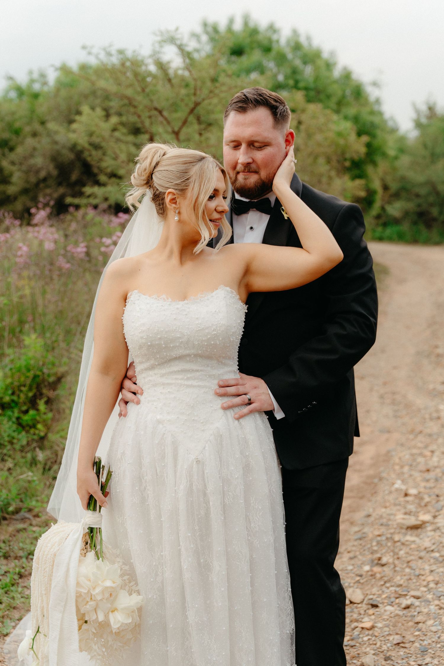 A romantic wedding couple embraces on a gravel path surrounded by blooming wildflowers and greenery.