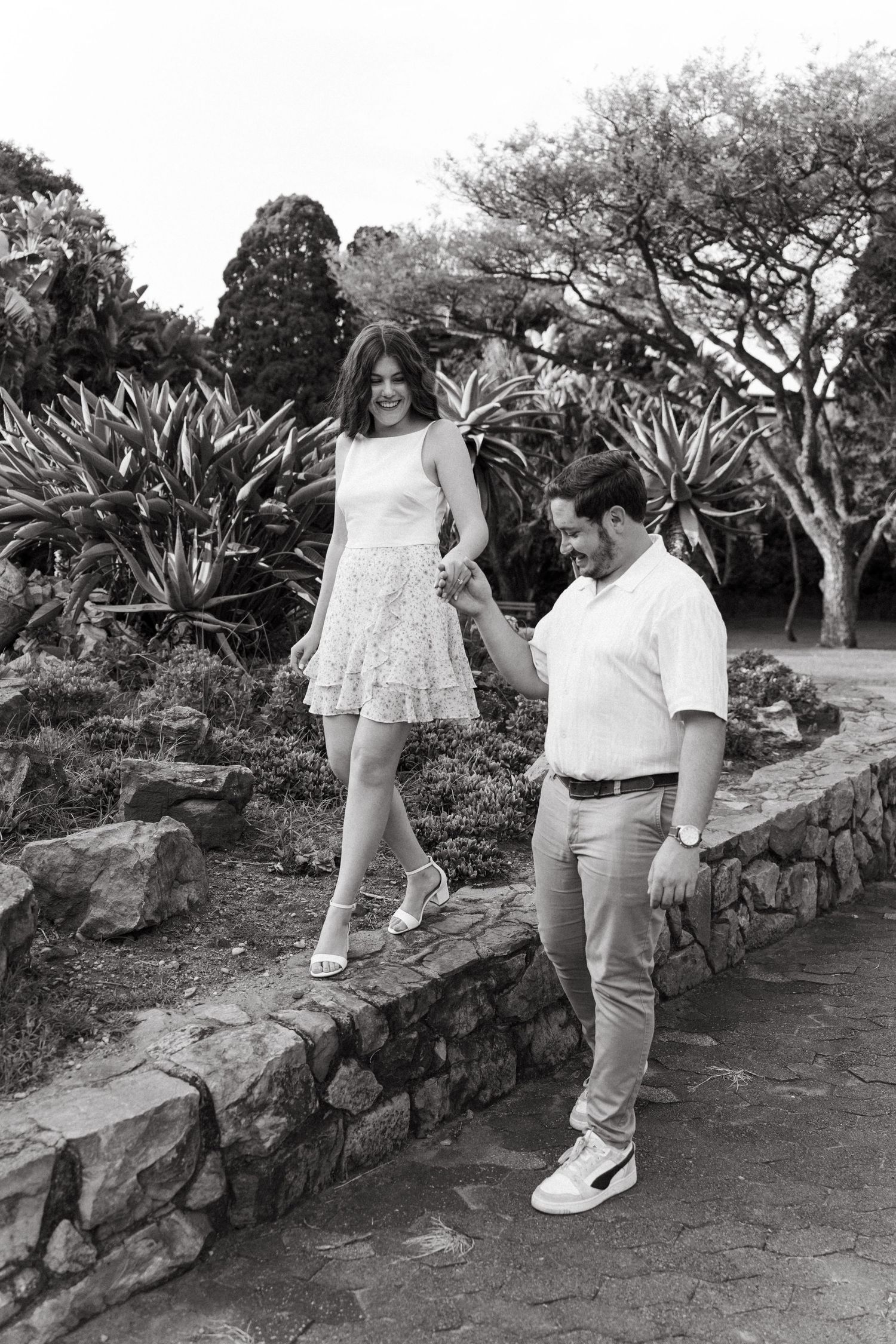 Black and white photo of a couple holding hands walking in a lush garden park, woman in dress balancing on stone wall.