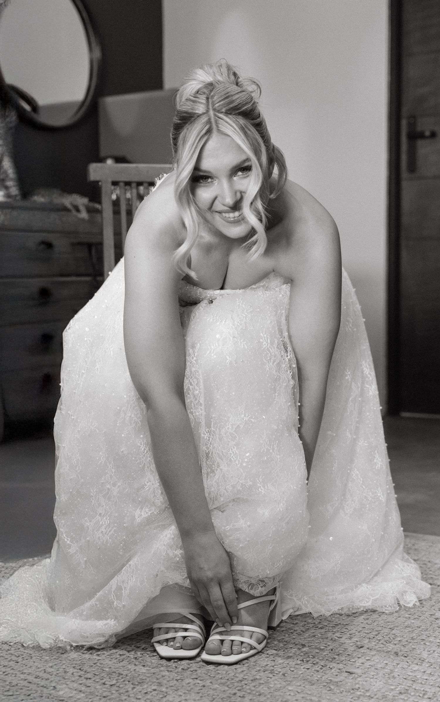 A bride in a white lace wedding dress adjusts her shoe while sitting on a carpeted floor.