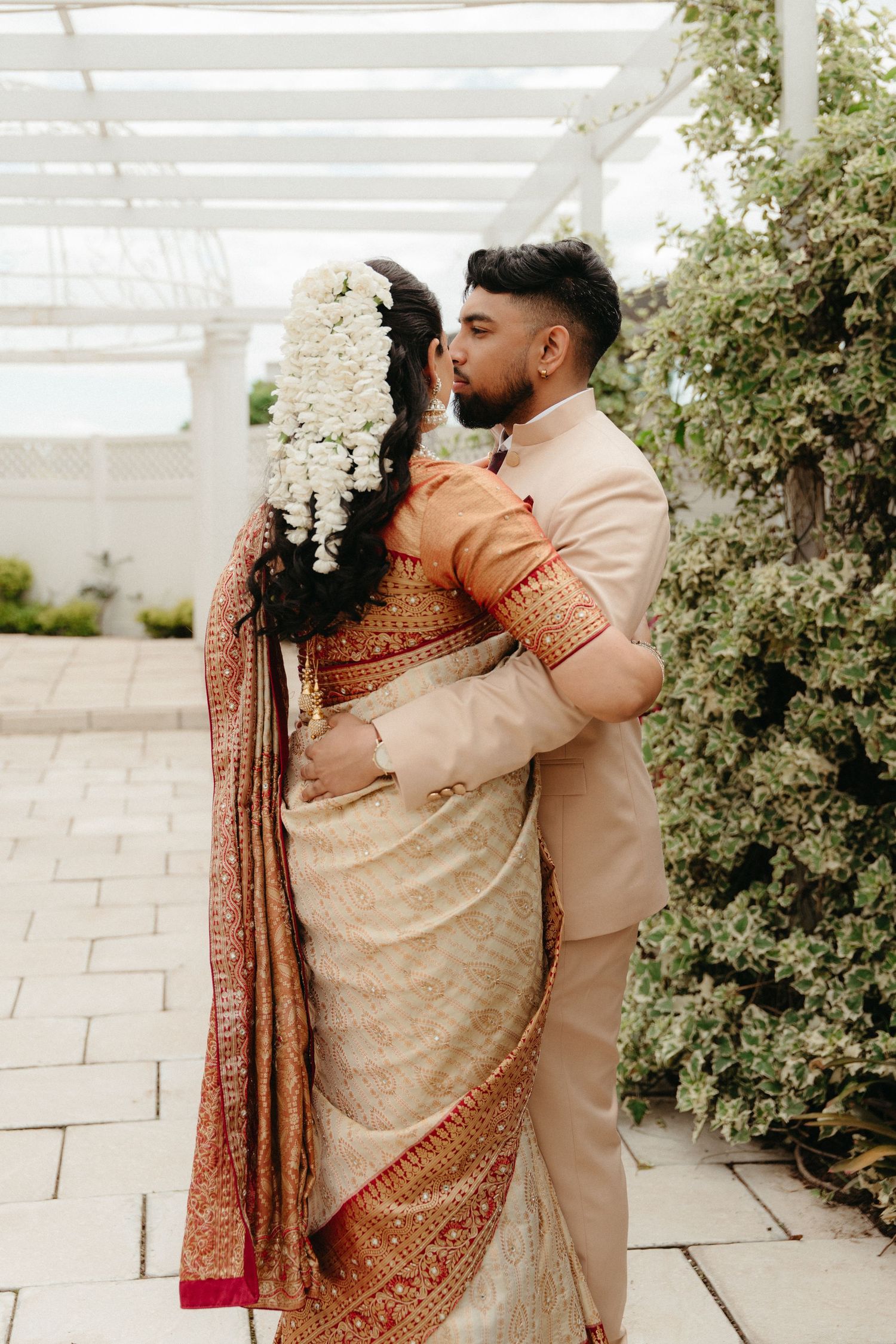 A couple in traditional Indian wedding attire embrace in a romantic moment against a white pergola and greenery backdrop.
