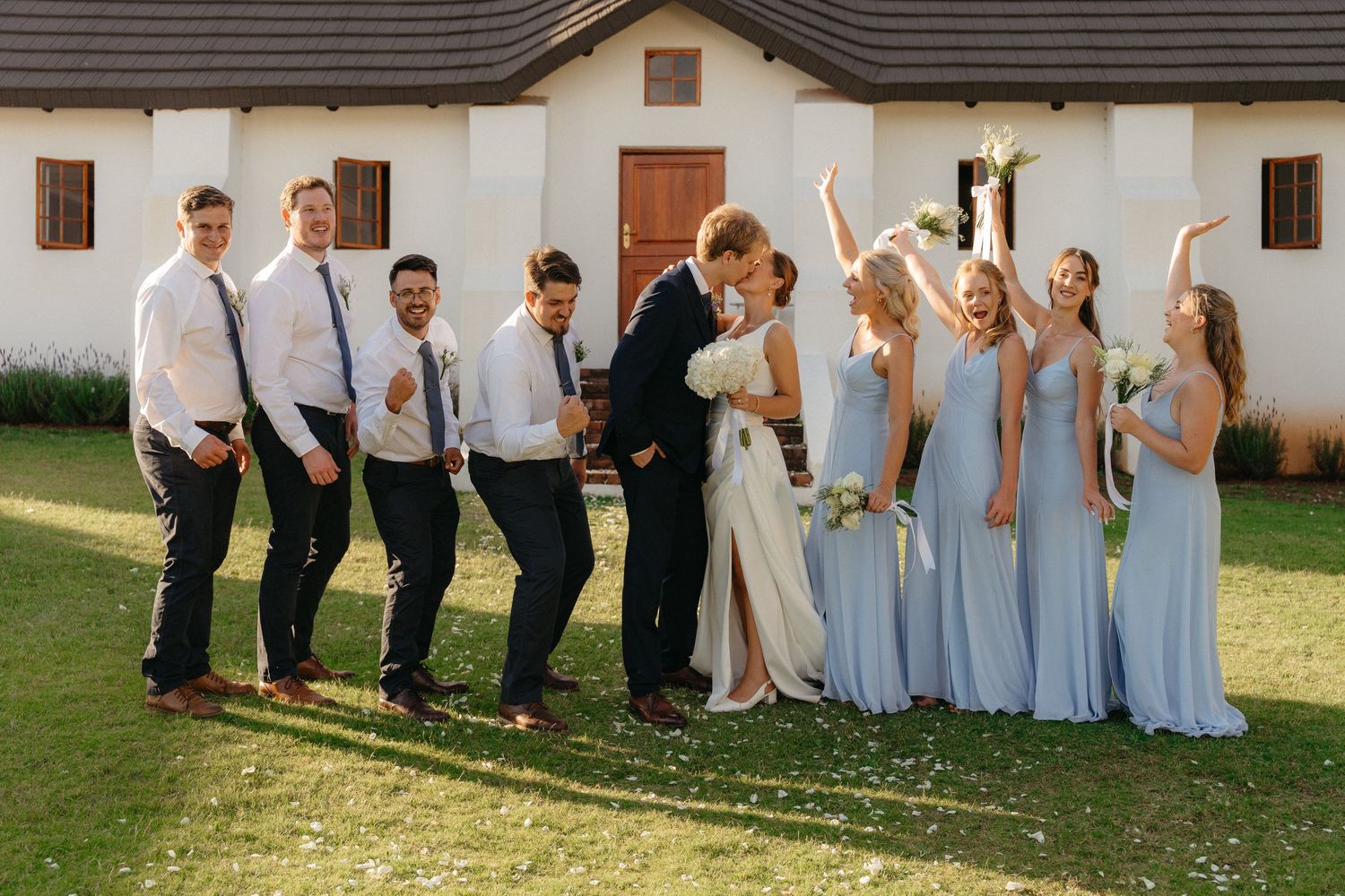 Wedding party poses creatively for fun group photos in light blue and white attire outside a rustic venue.