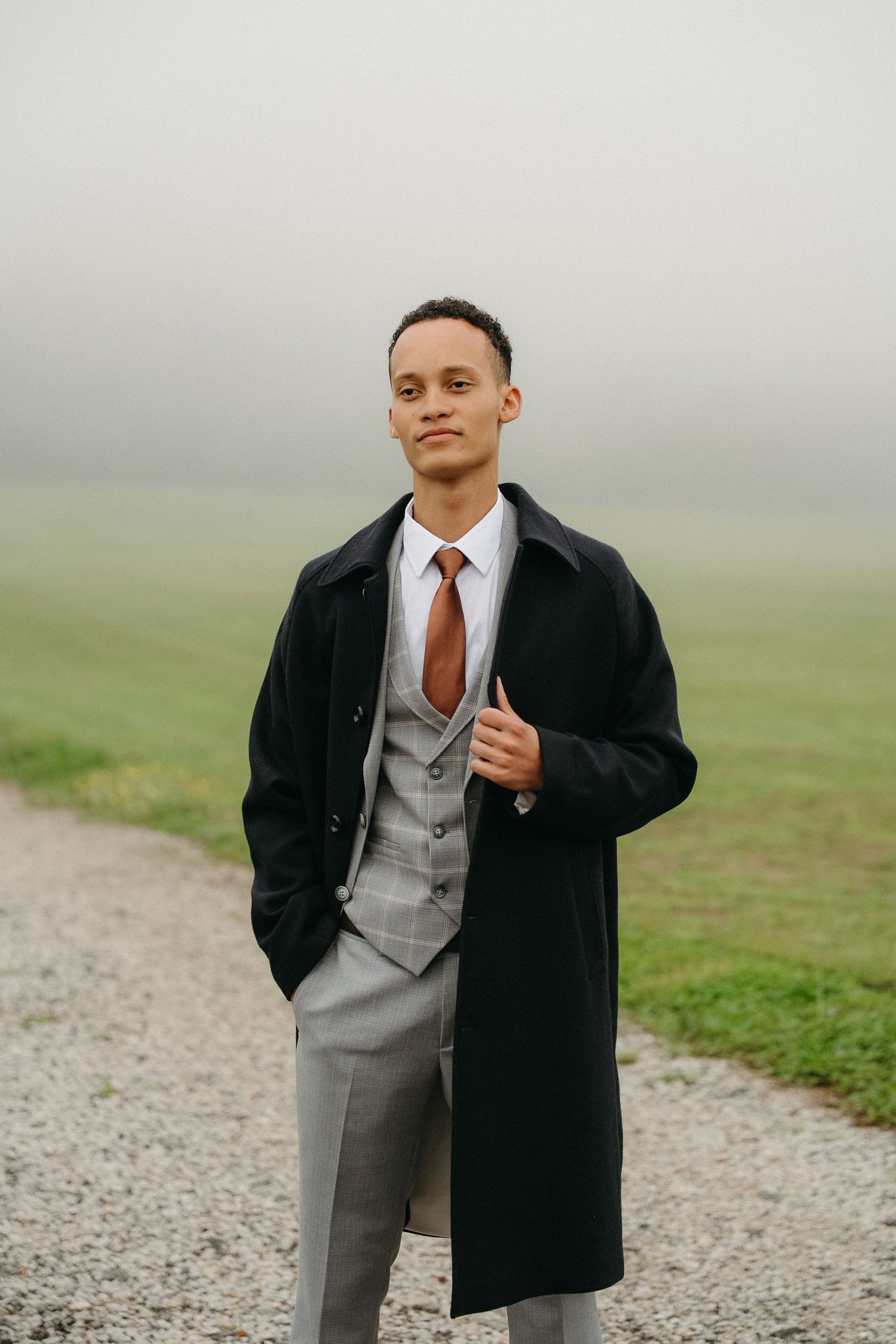 A groom in a black overcoat, gray suit, and brown tie stands on a gravel path in a misty green field.