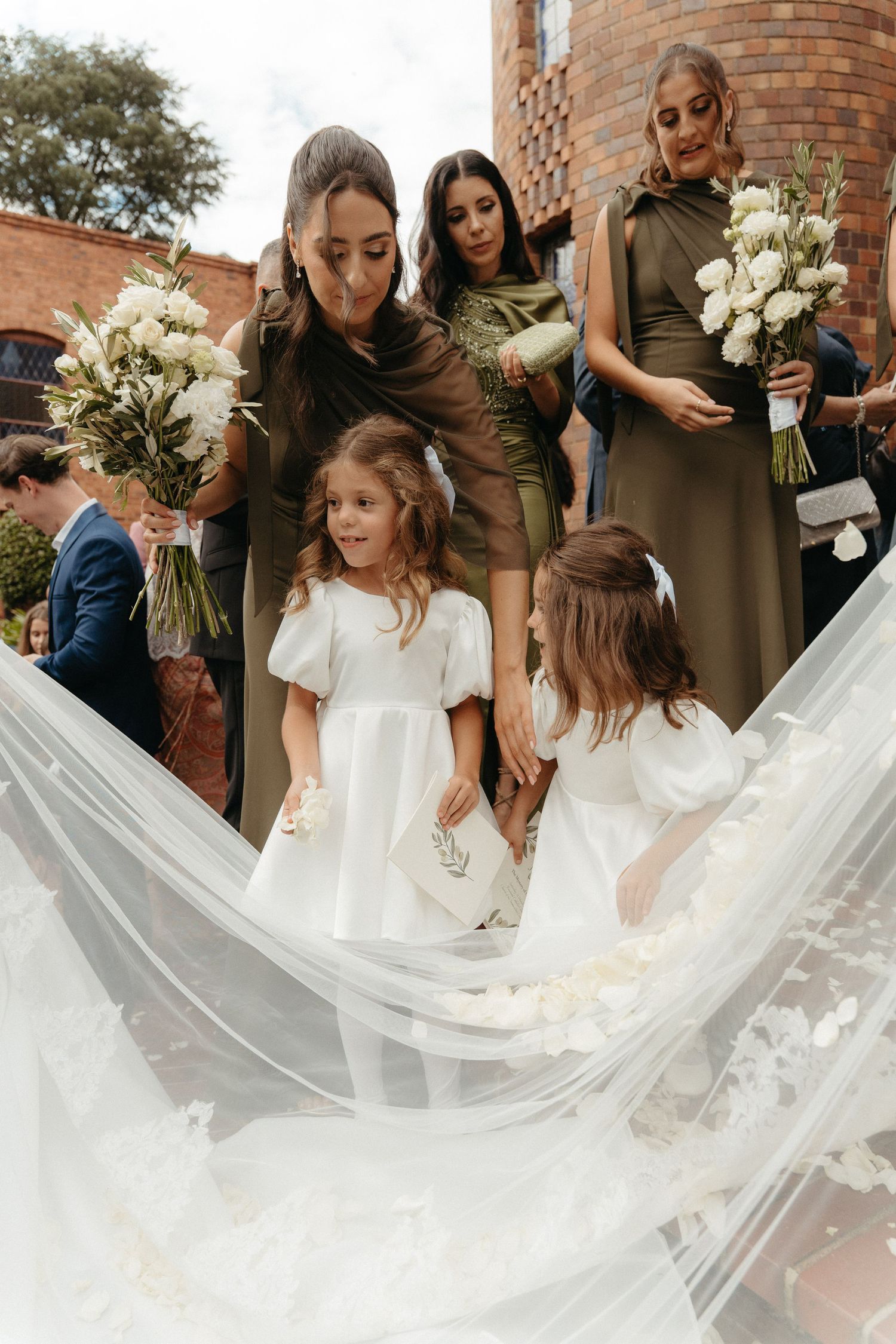 Bridesmaids in olive dresses hold white bouquets as flower girls in white dresses arrange a bridal veil outdoors.