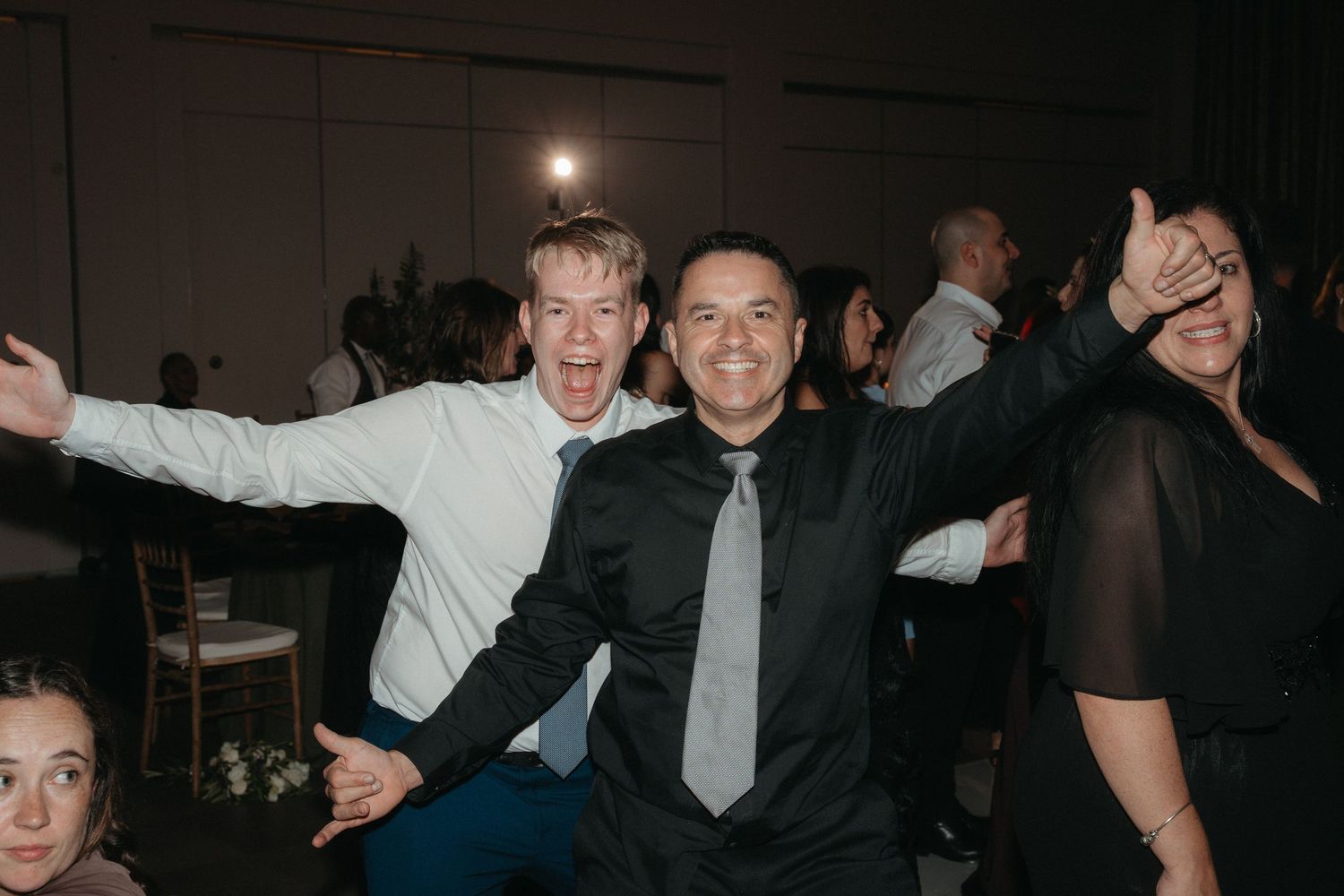 Two joyful men celebrating at a wedding reception, arms outstretched, smiling broadly in formal attire.
