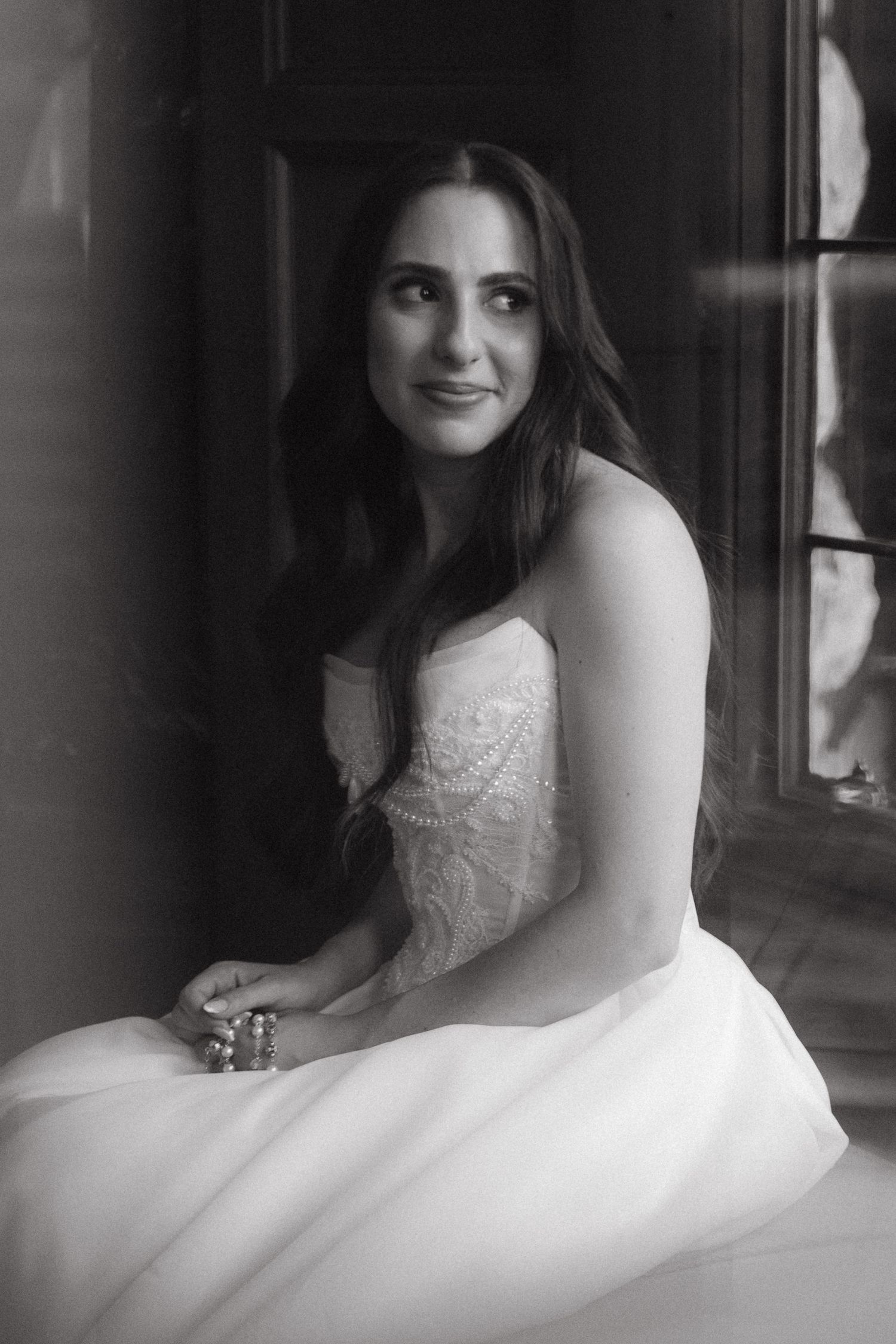 Black and white portrait of a bride in a strapless lace gown sitting by a window, smiling softly.