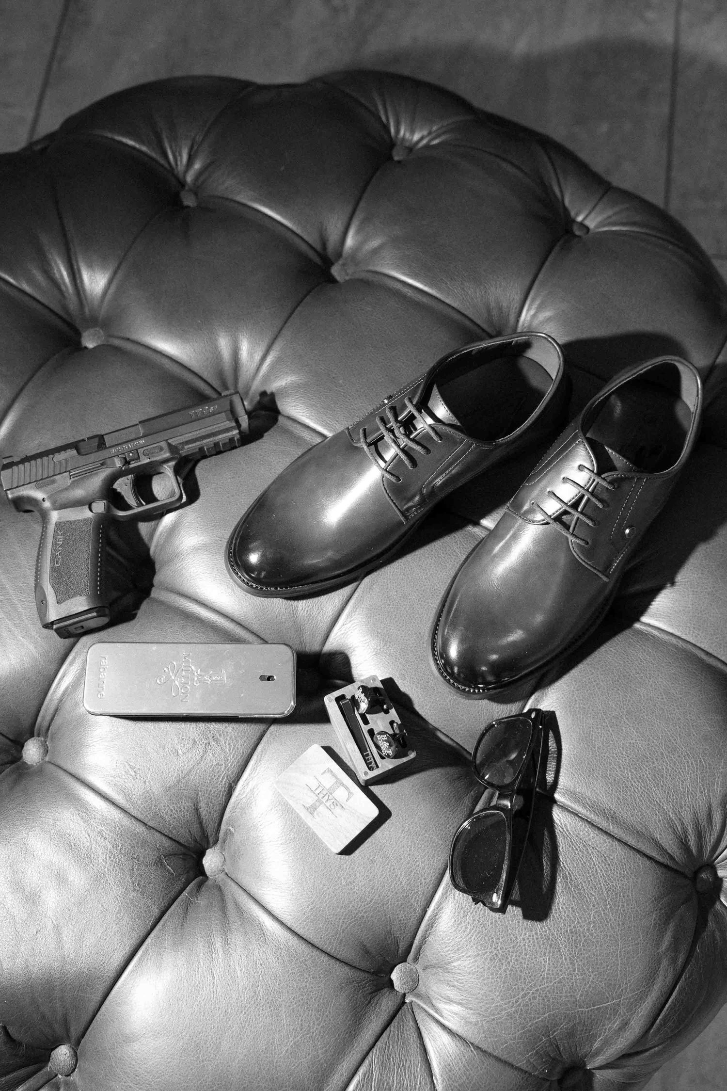 Black and white flat lay of mens dress shoes, handgun, sunglasses, lighter, and phone on tufted leather surface.