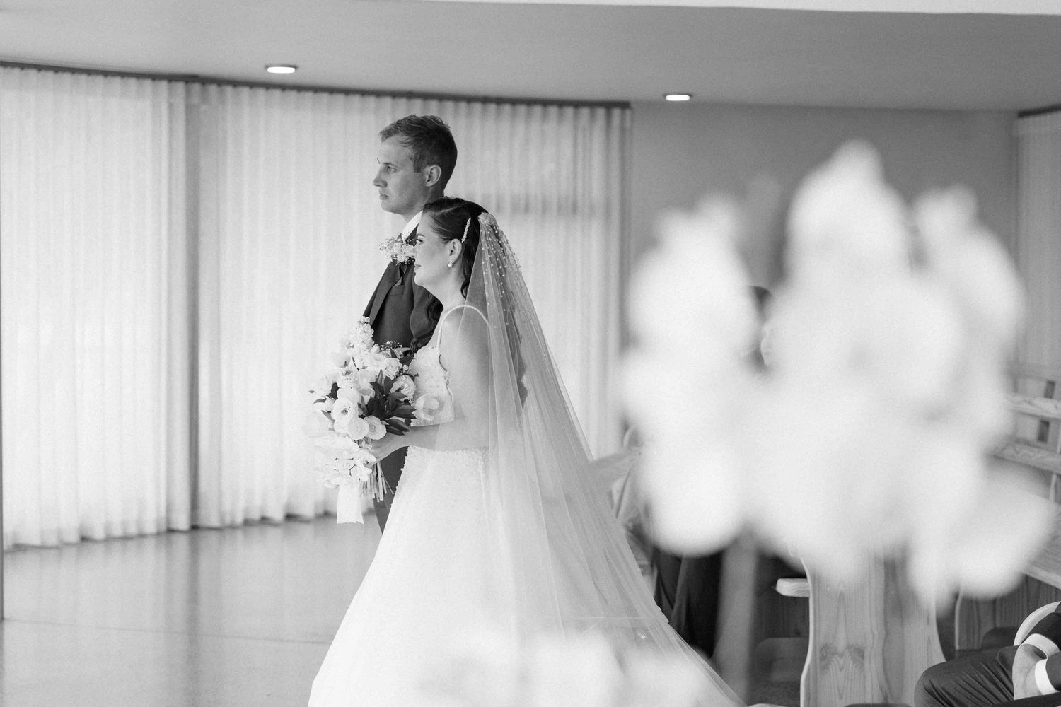 Black and white wedding photo of a bride in a flowing gown and veil holding a bouquet indoors.
