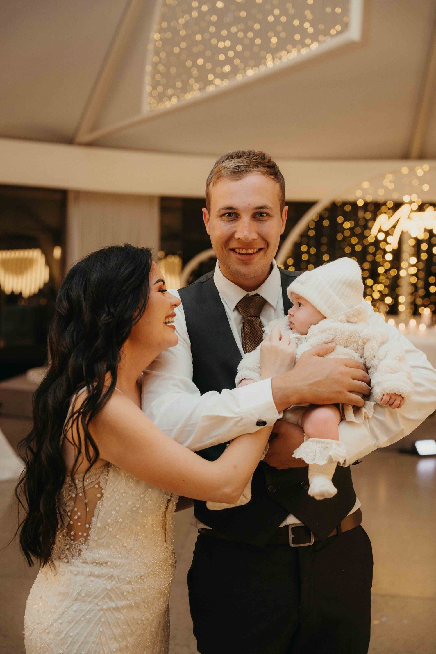 A smiling couple in formal wedding attire holds their baby dressed in white at an elegant reception venue.