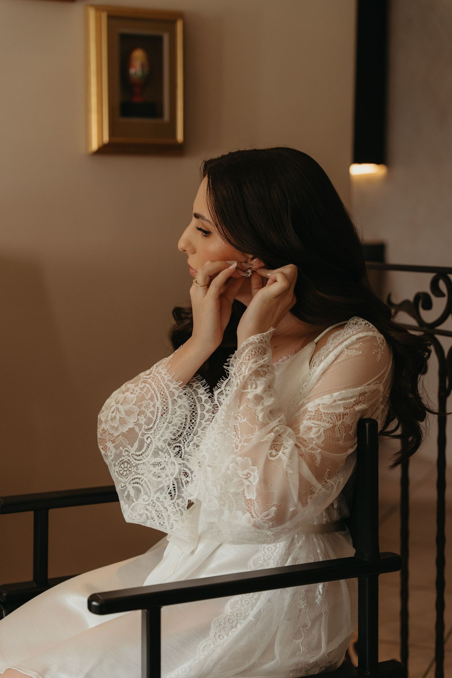 Bride in white lace gown putting on earrings while seated on ornate black chair in elegant room  in Johannesburg.