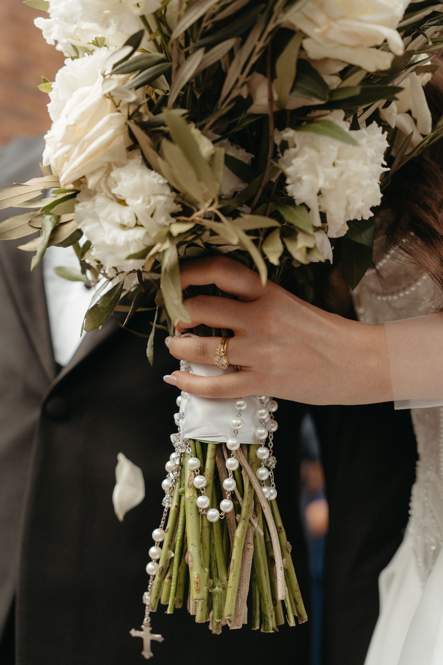 Bride holding white peony wedding bouquet with rosary beads and cross, wearing gold ring, beside groom in gray suit.