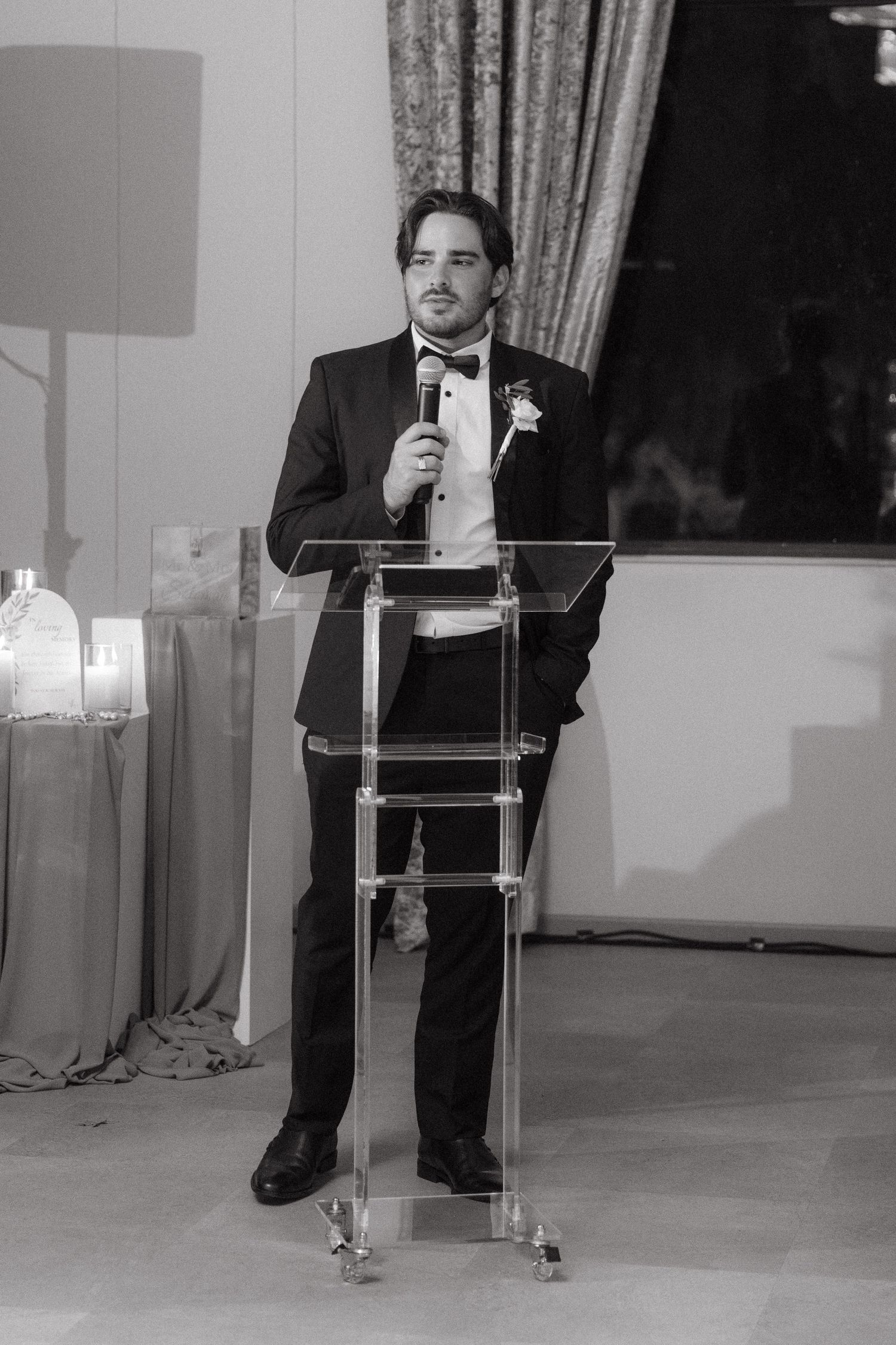 Man in tuxedo with boutonniere speaks at clear acrylic podium with microphone at formal wedding reception.