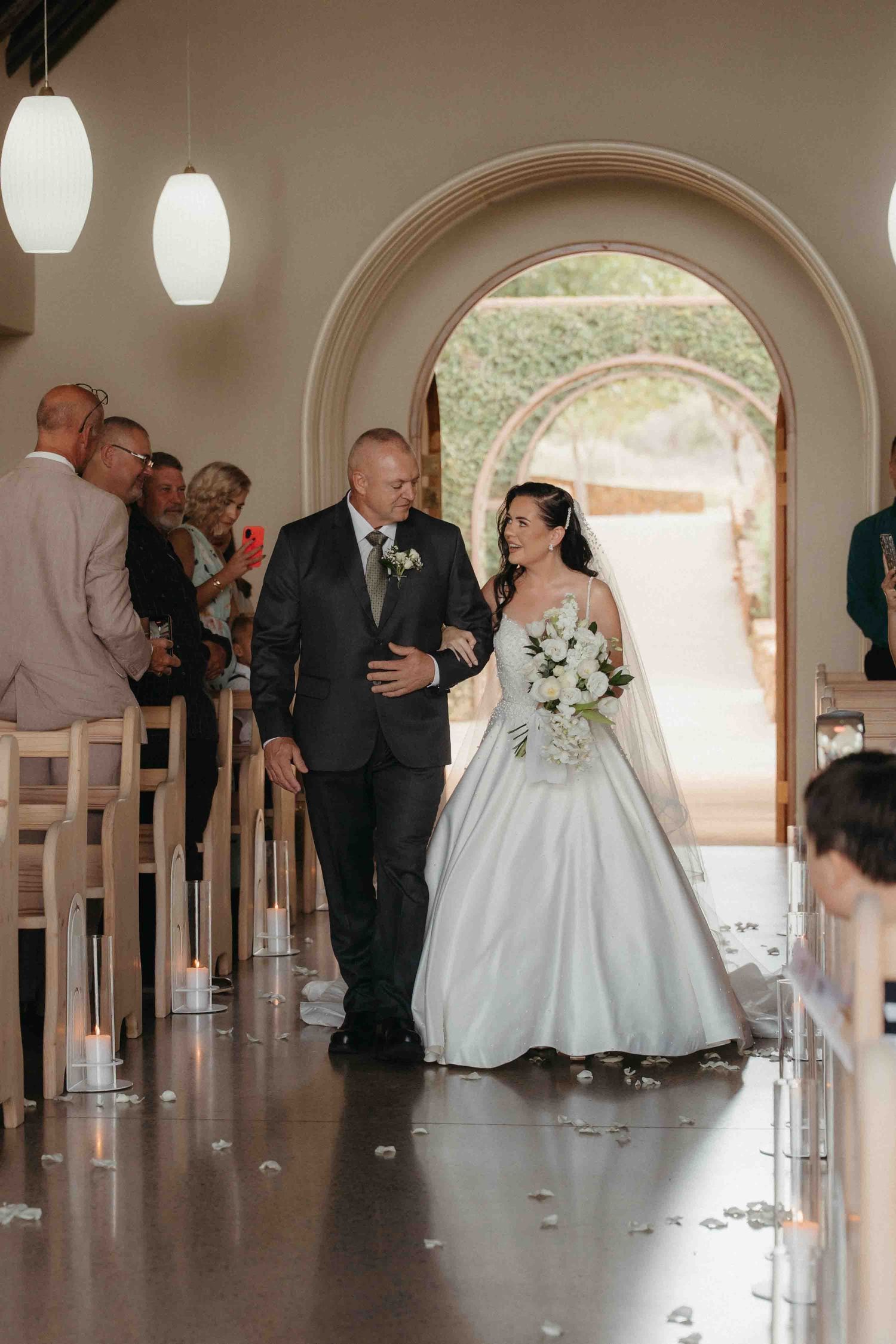 Bride in white ball gown walks down church aisle with escort, holding white bouquet, guests seated in pews.