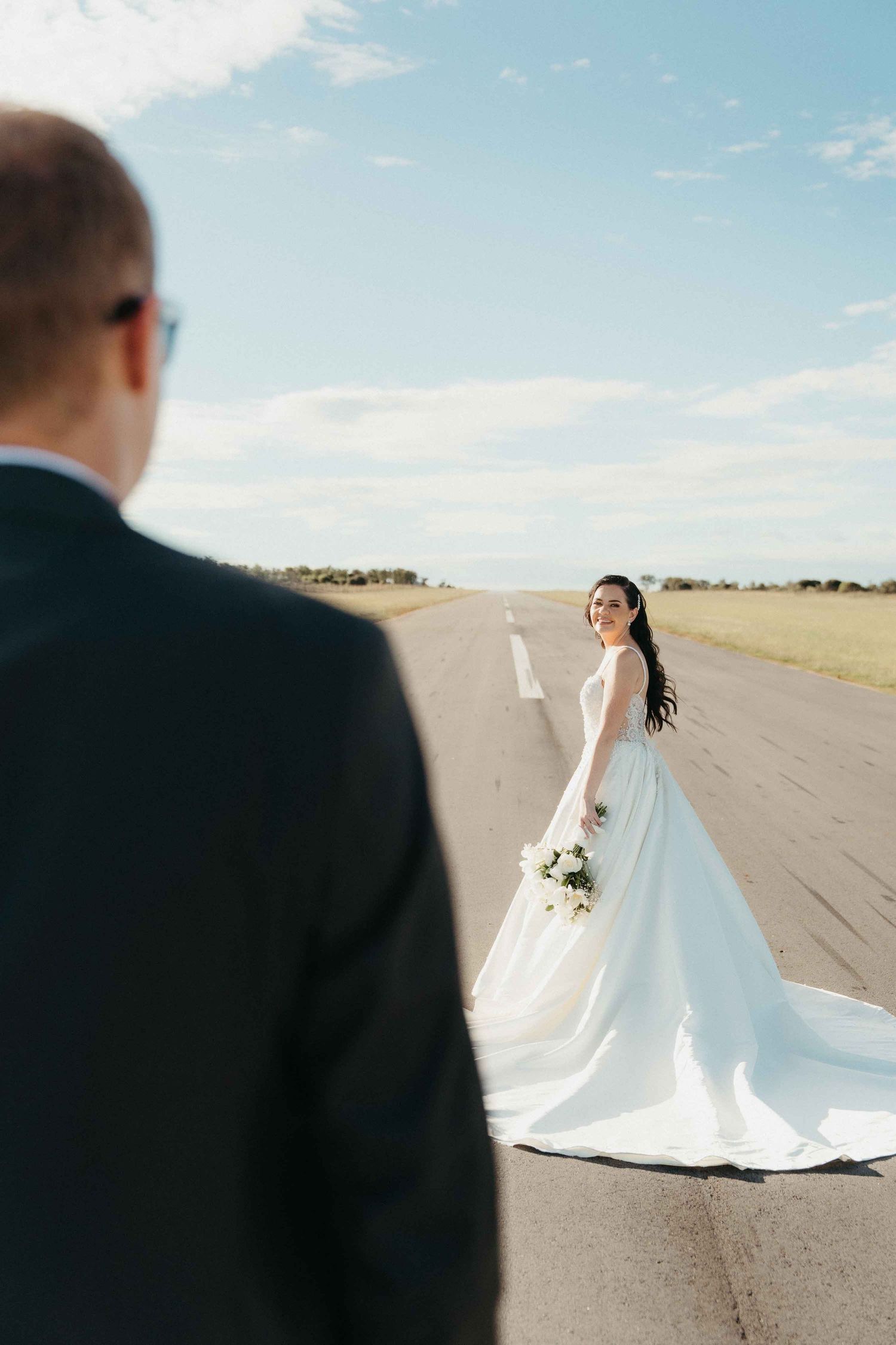 Bride in white gown holds bouquet on runway, smiling back at groom in dark suit under blue sky.