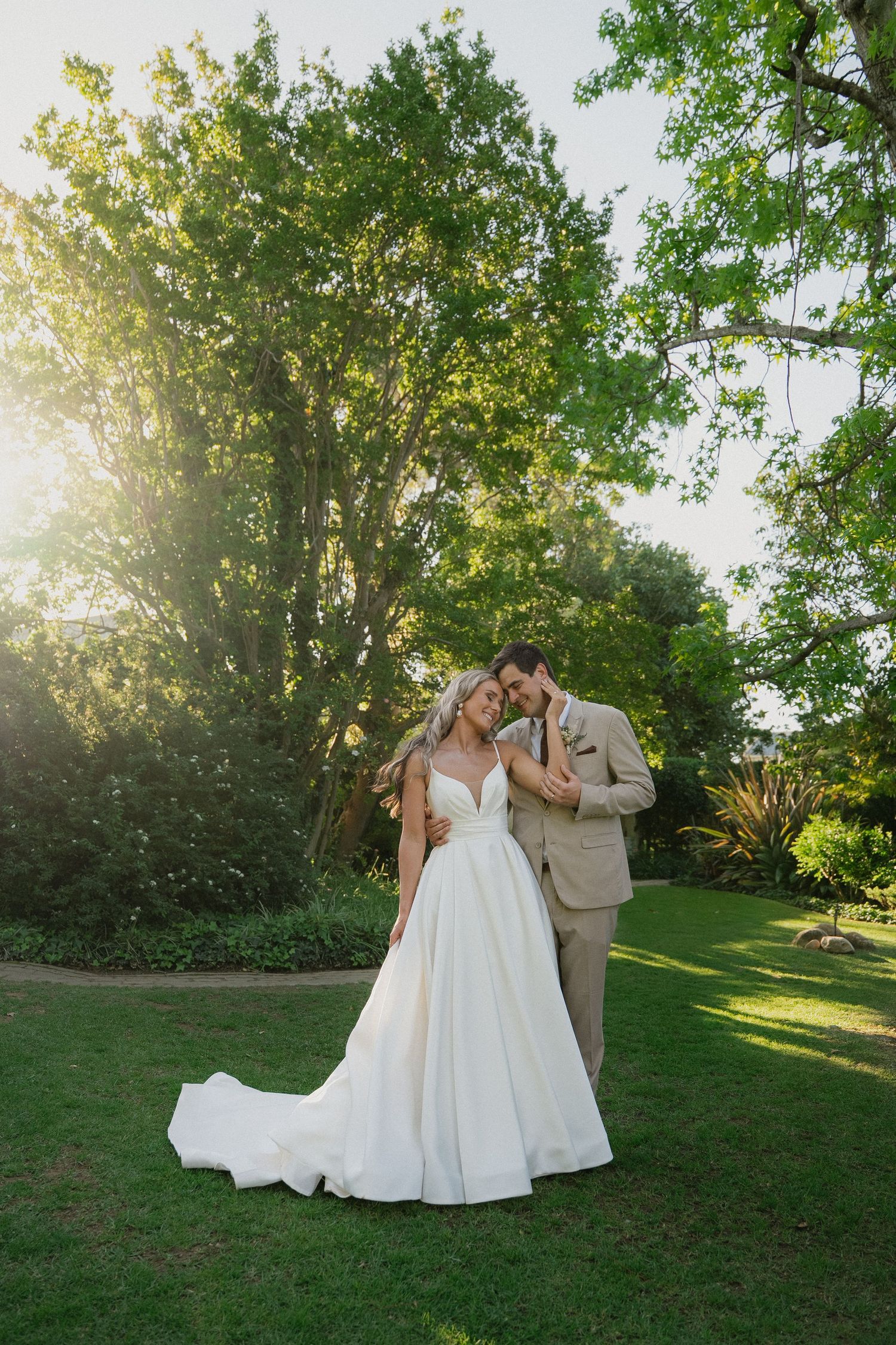 Bride in white ball gown and groom in tan suit embrace in lush garden with sunlight filtering through trees.