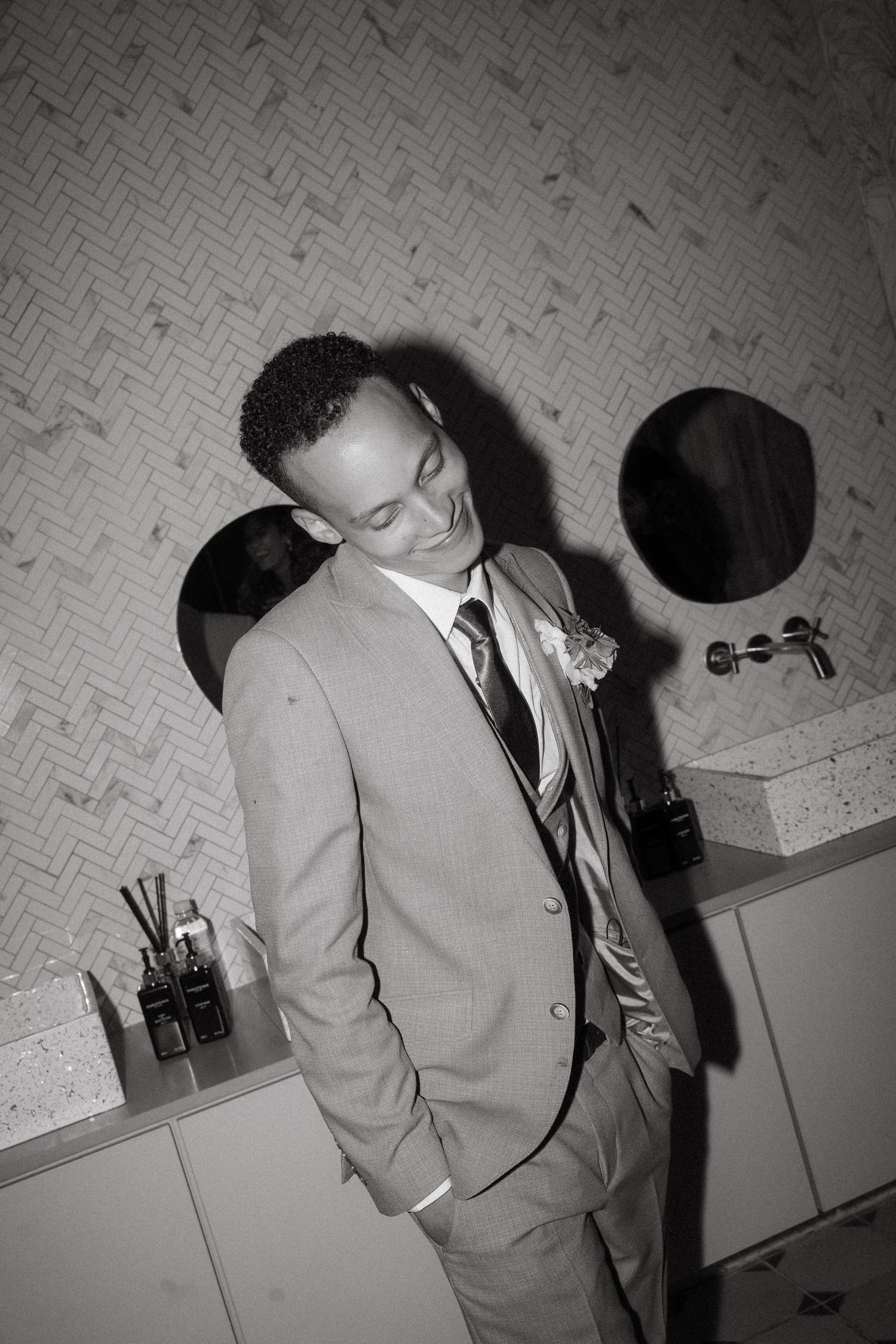 Black and white photo of a groom in a suit and tie with a boutonniere, standing in a modern bathroom.