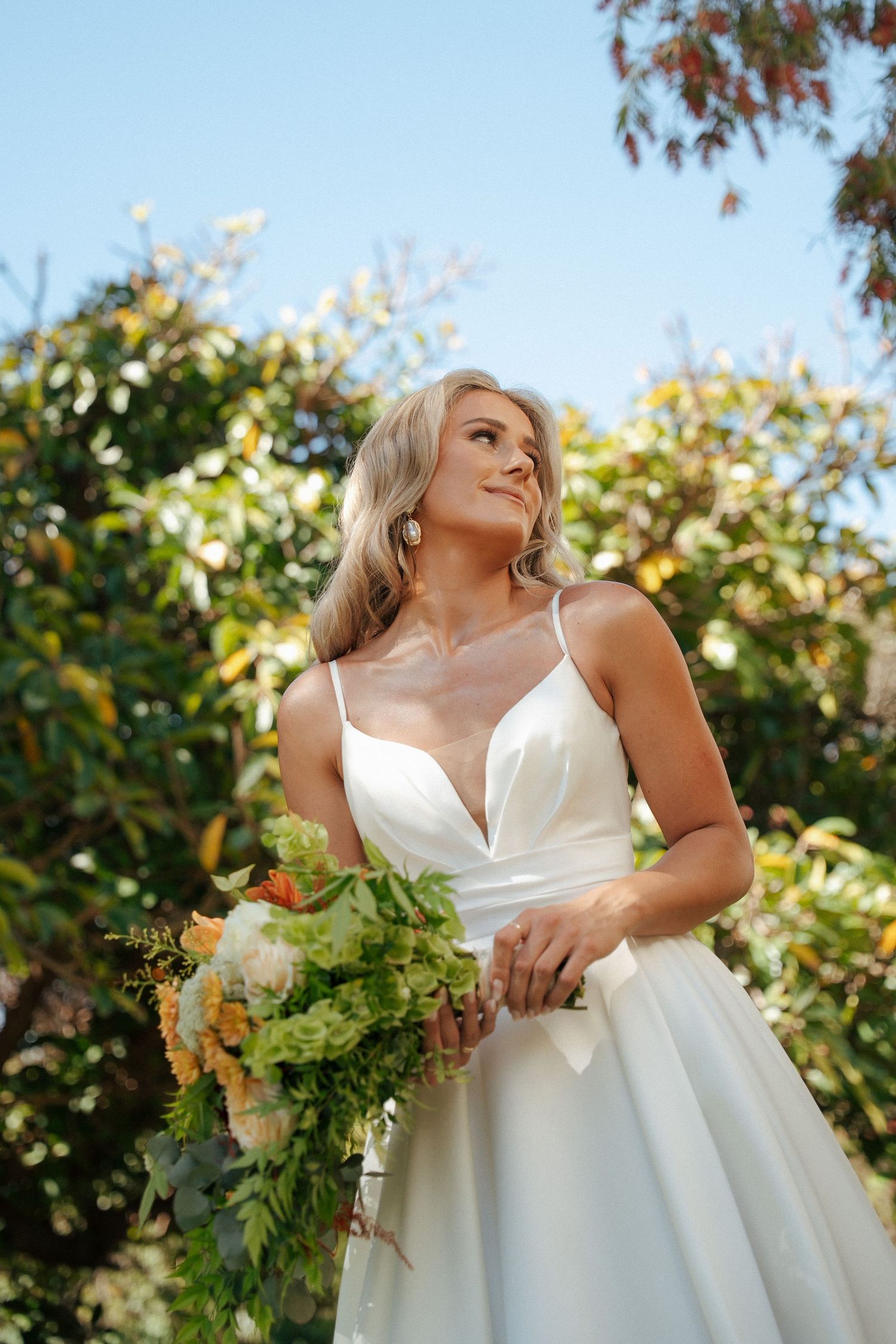 Smiling bride in a white V-neck satin wedding gown holds a colorful bouquet of orange and green flowers outdoors beneath a clear blue sky.