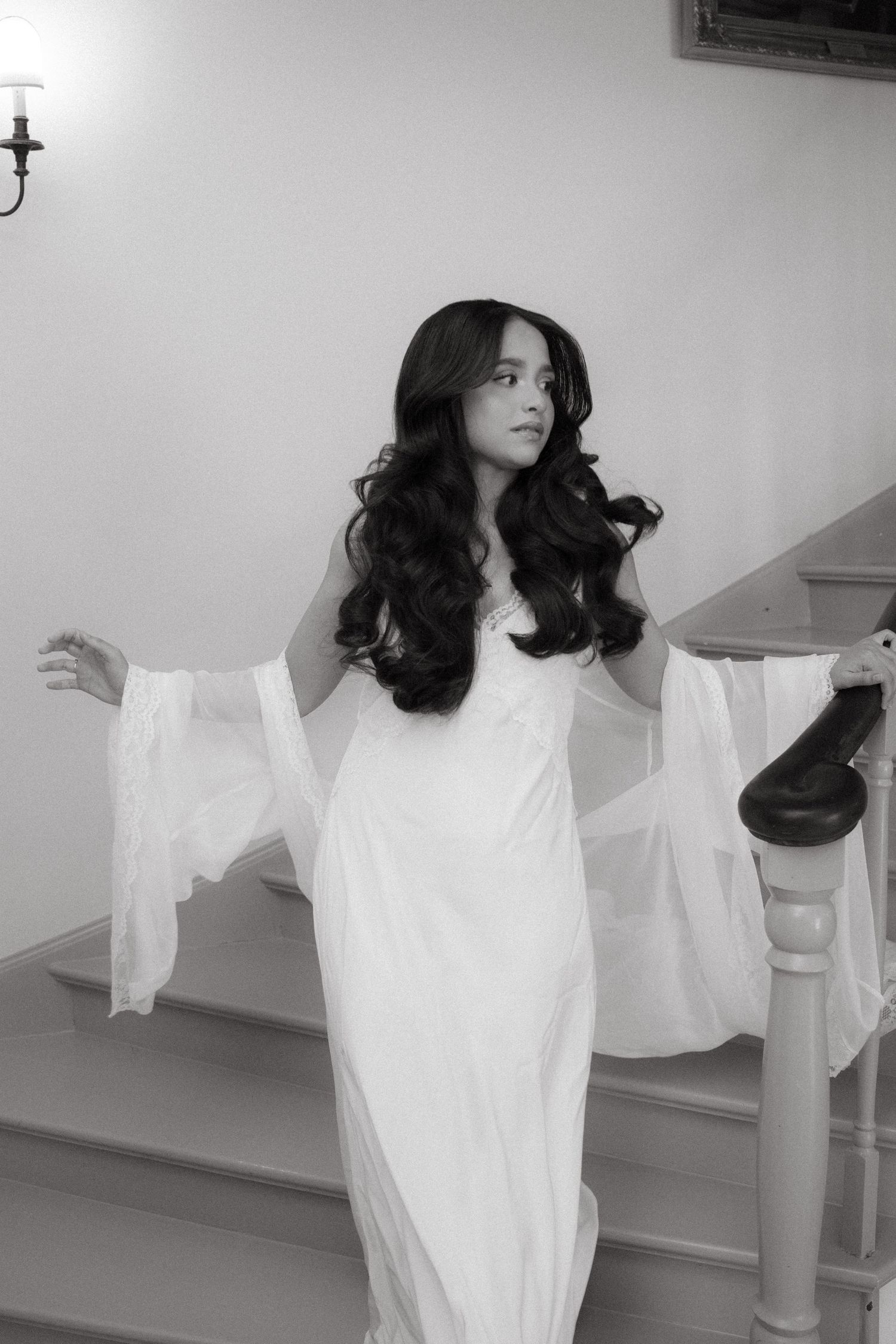Bride with long curly hair in white dress with flowing sleeves poses on staircase at Beechwood Gardens, black and white.