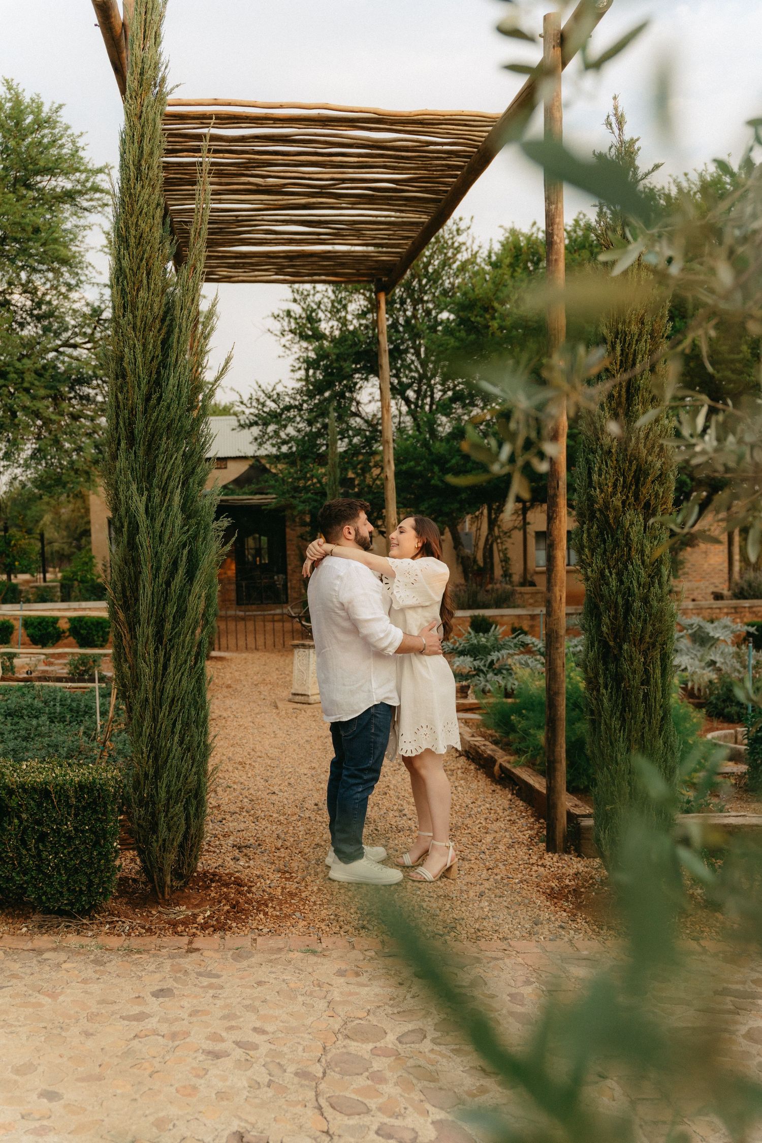 Couple sharing romantic kiss under bamboo pergola at Campagne Living surrounded by cypress trees in garden setting.